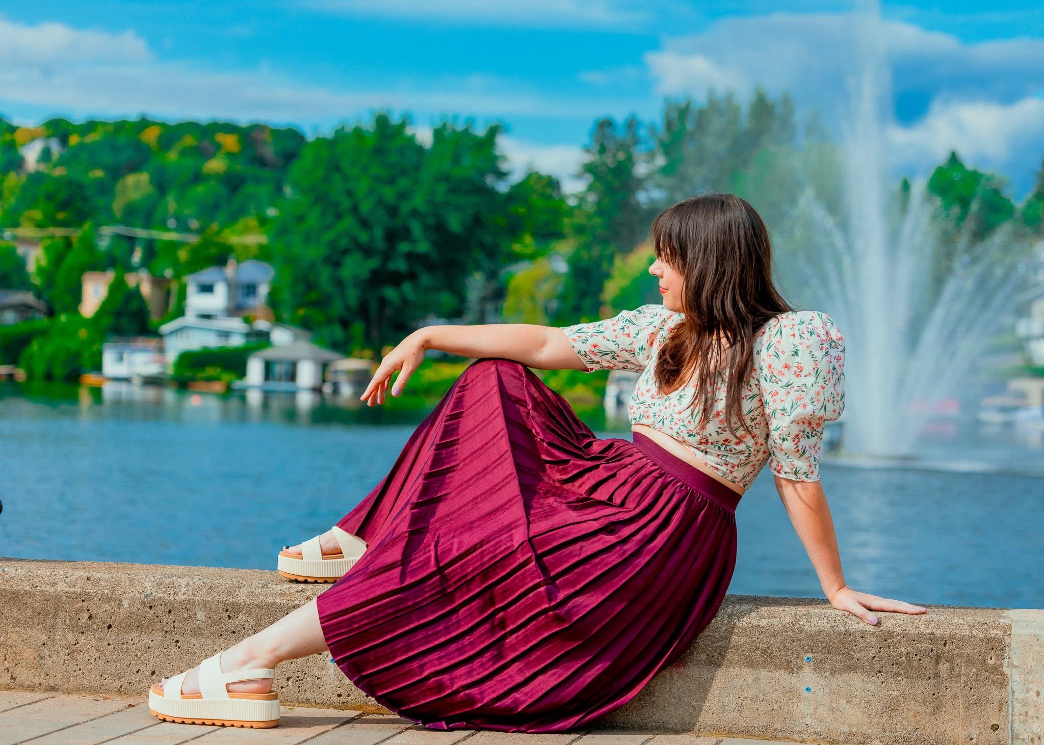 A woman sitting on a concrete ledge near a lake, with a fountain and trees in the background, wearing a floral top and a pleated maroon skirt.