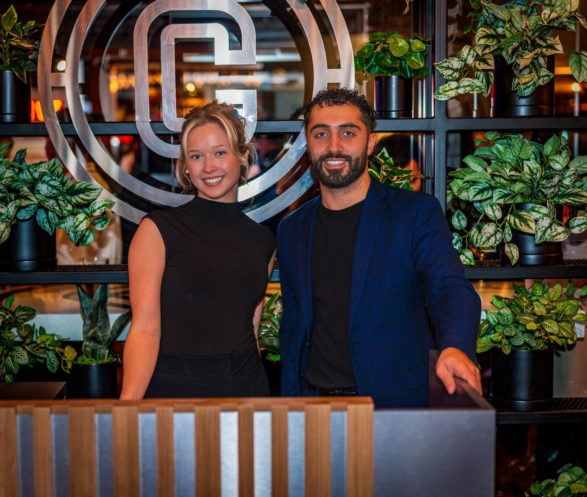A young woman and man standing behind a reception desk at a restaurant, smiling, with greenery and a logo behind them.