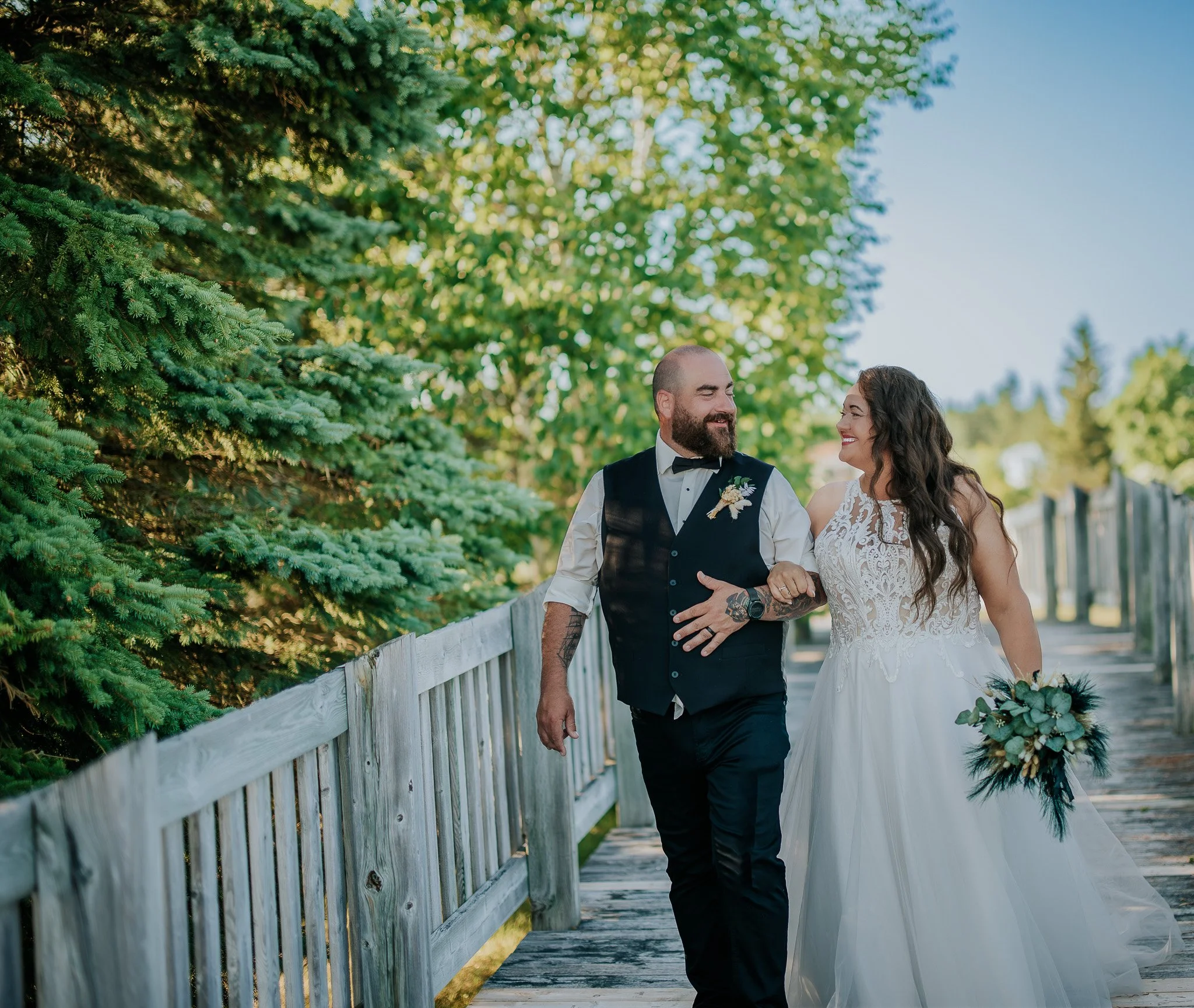 A bride and a groom walk arm in arm on a wooden pathway surrounded by green trees. The groom is dressed in a black vest, white shirt, and black pants, while the bride is wearing a white wedding gown and holding a bouquet of greenery and flowers. They