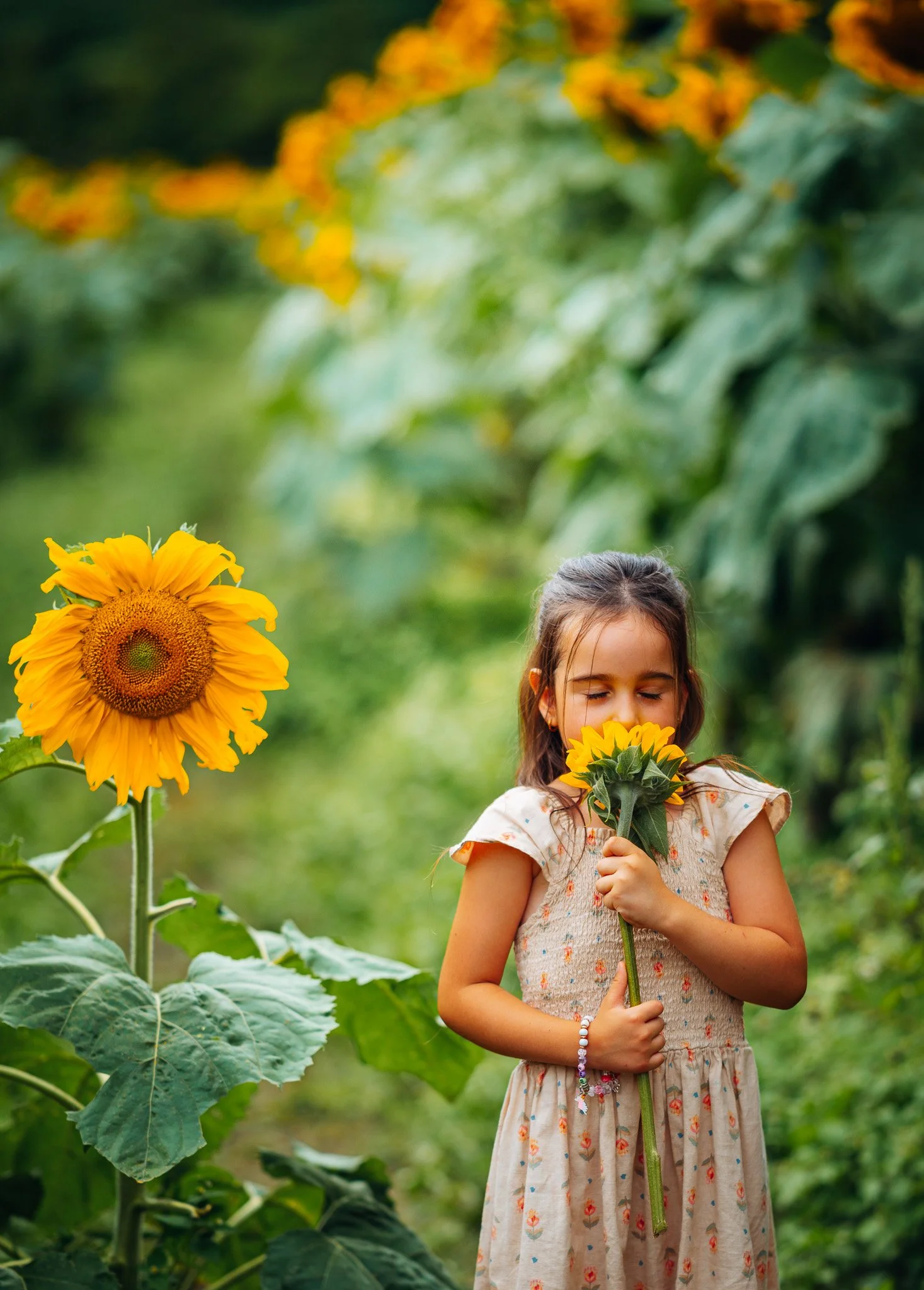 A young girl with brown hair and closed eyes holding a sunflower stem in a garden with large green leaves and sunflower blooms.
