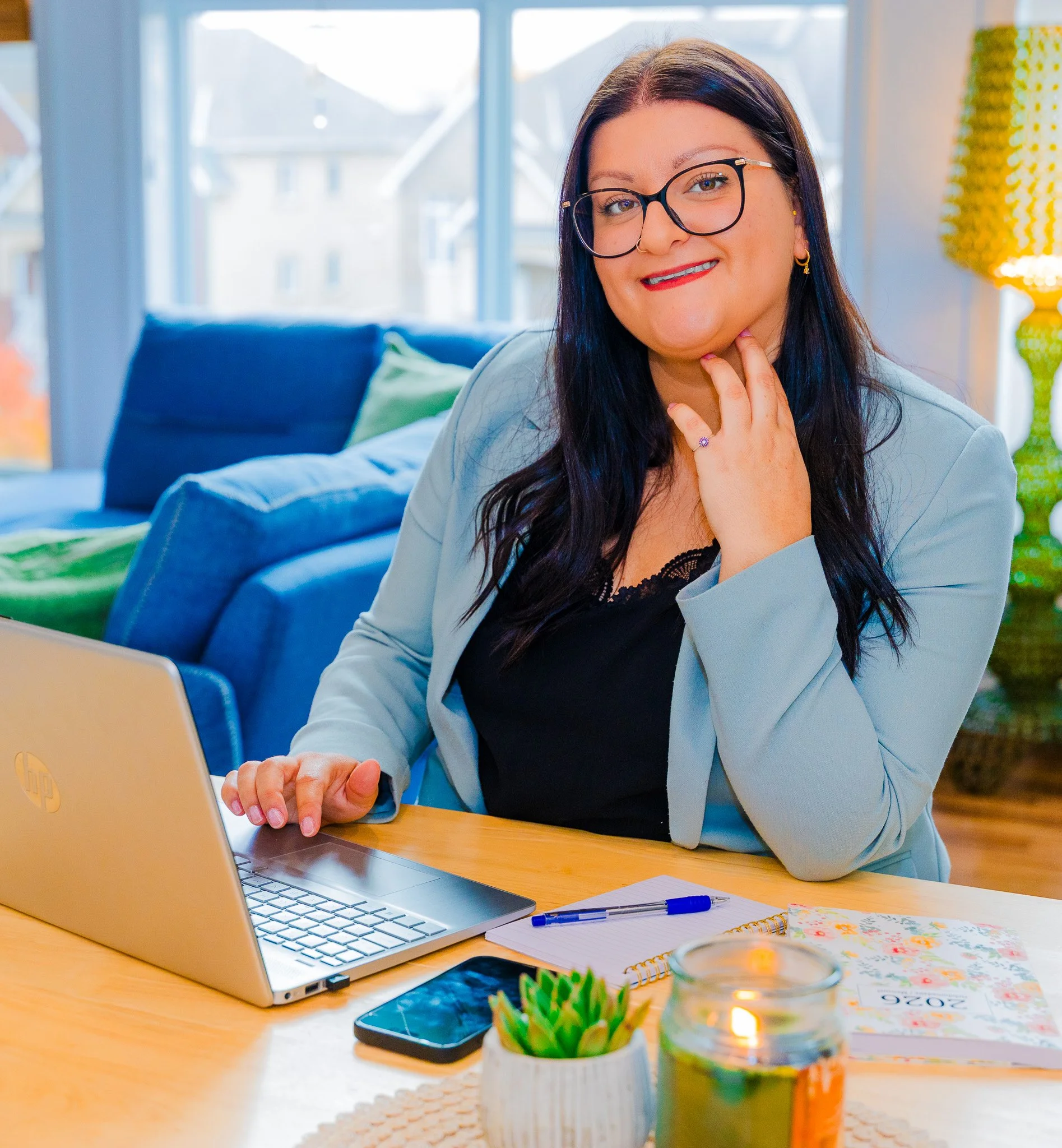 A woman with dark hair and glasses sitting at a wooden desk, using a silver laptop. She has a ring on her finger, a notepad, a pen, a smartphone, a small potted plant, and a candle on the desk. In the background, large windows show a residential neig