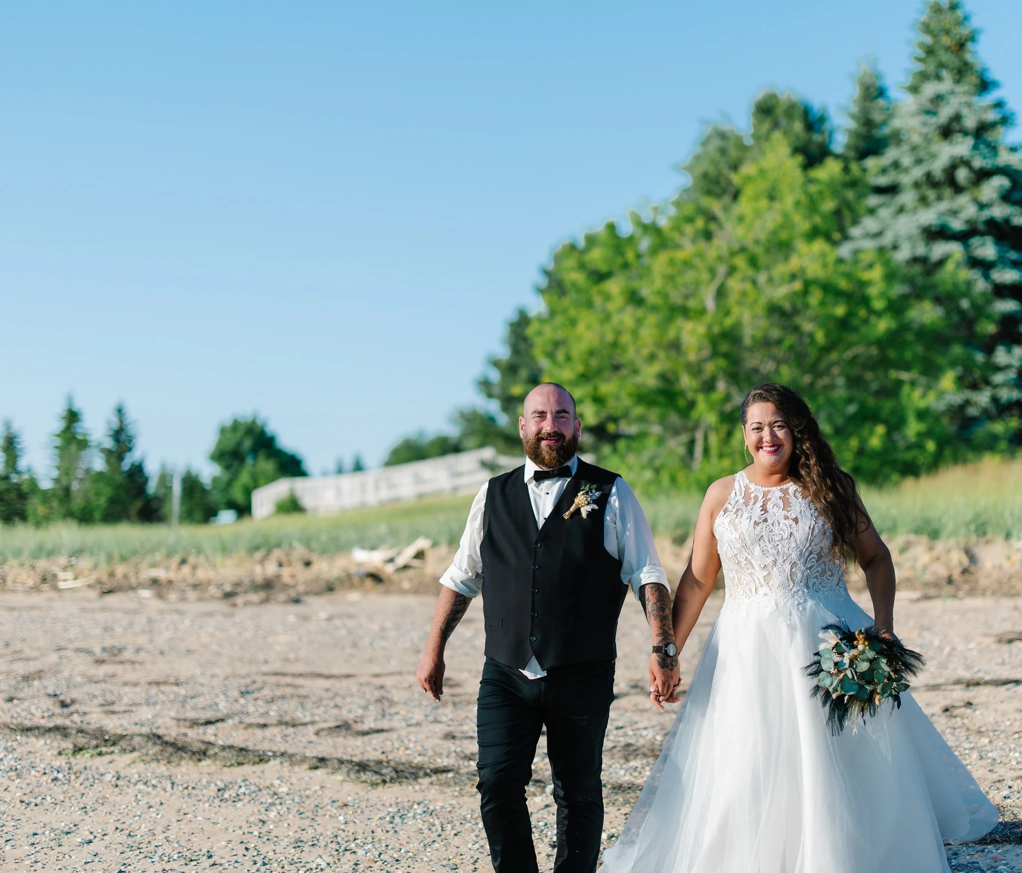 A newlywed couple walking hand in hand outdoors on a sunny day, with green trees and a blue sky in the background, carrying a bouquet of flowers.