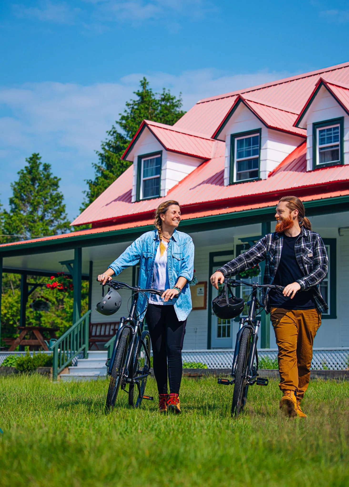 A young man and woman walking with bicycles in front of a house on a bright, sunny day.