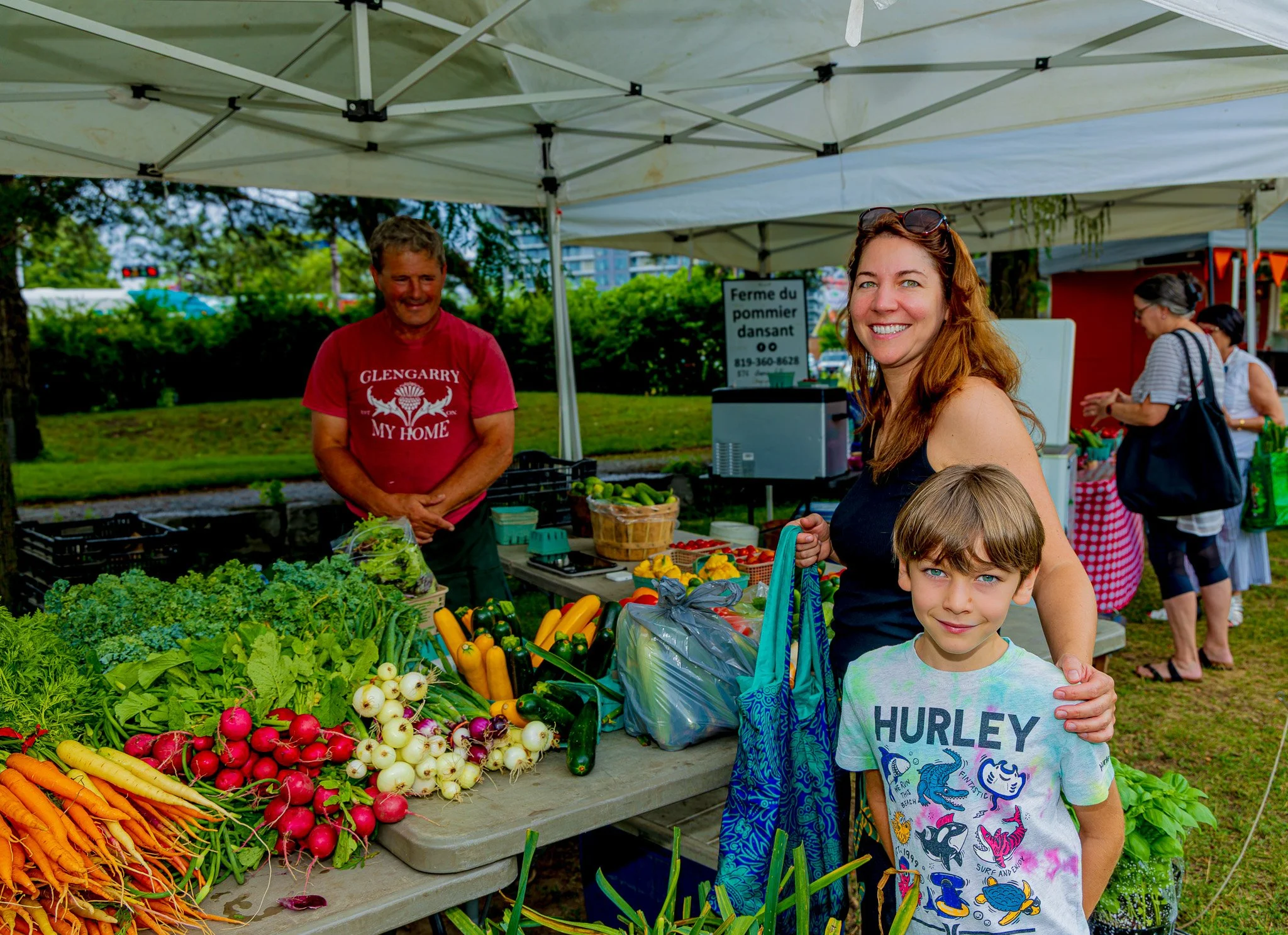 A woman and a boy shopping at a farmers market stall with fresh vegetables, with other shoppers in the background and a man selling produce.