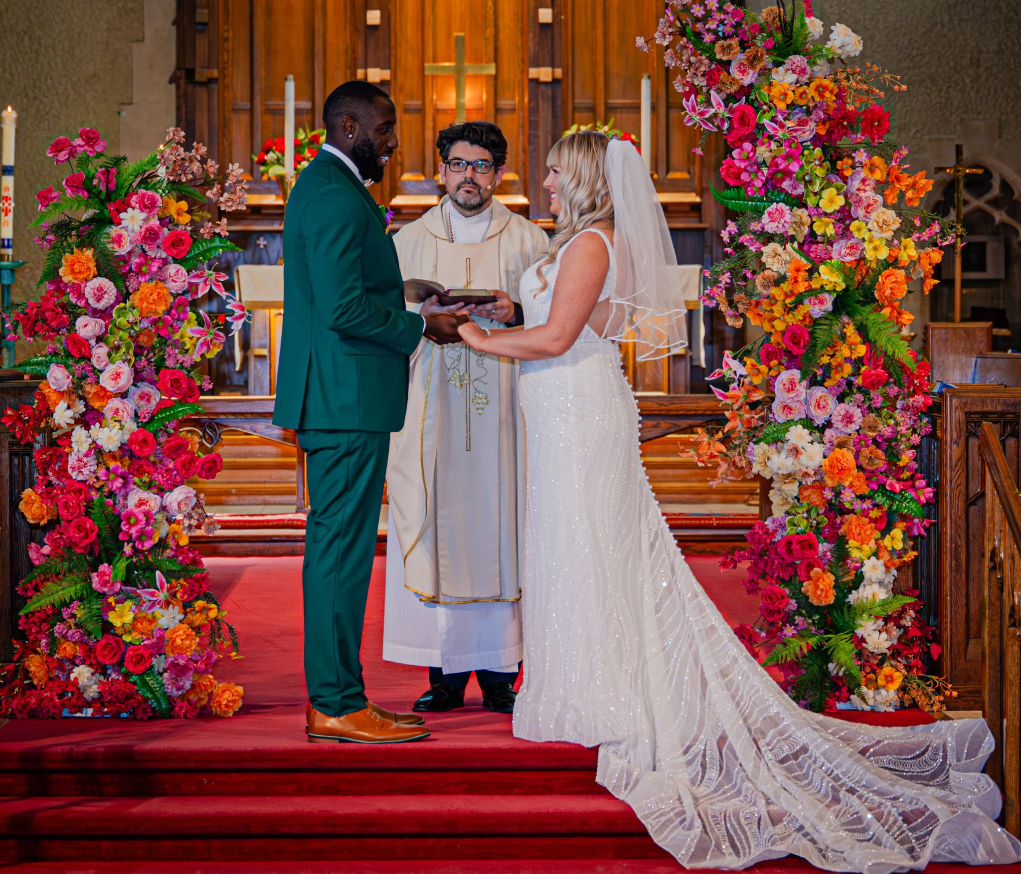 A wedding ceremony taking place in a church with the bride and groom exchanging vows, surrounded by colorful flower arrangements, with a priest officiating.