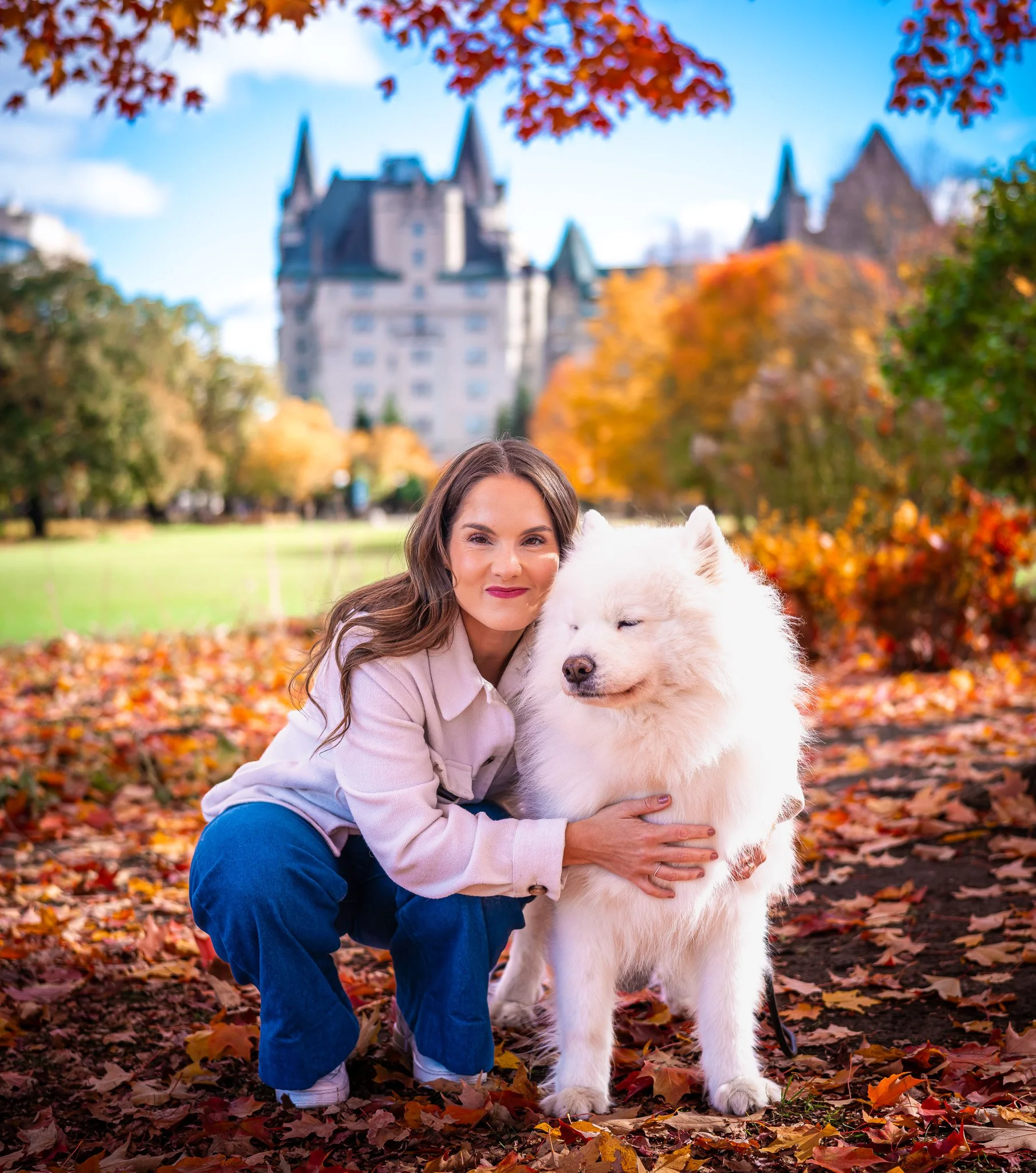 A woman with long brown hair smiling while hugging a large white dog with a fluffy coat and closed eyes, outdoors in a park with autumn leaves on the ground and colorful trees, and a castle-like building in the background.