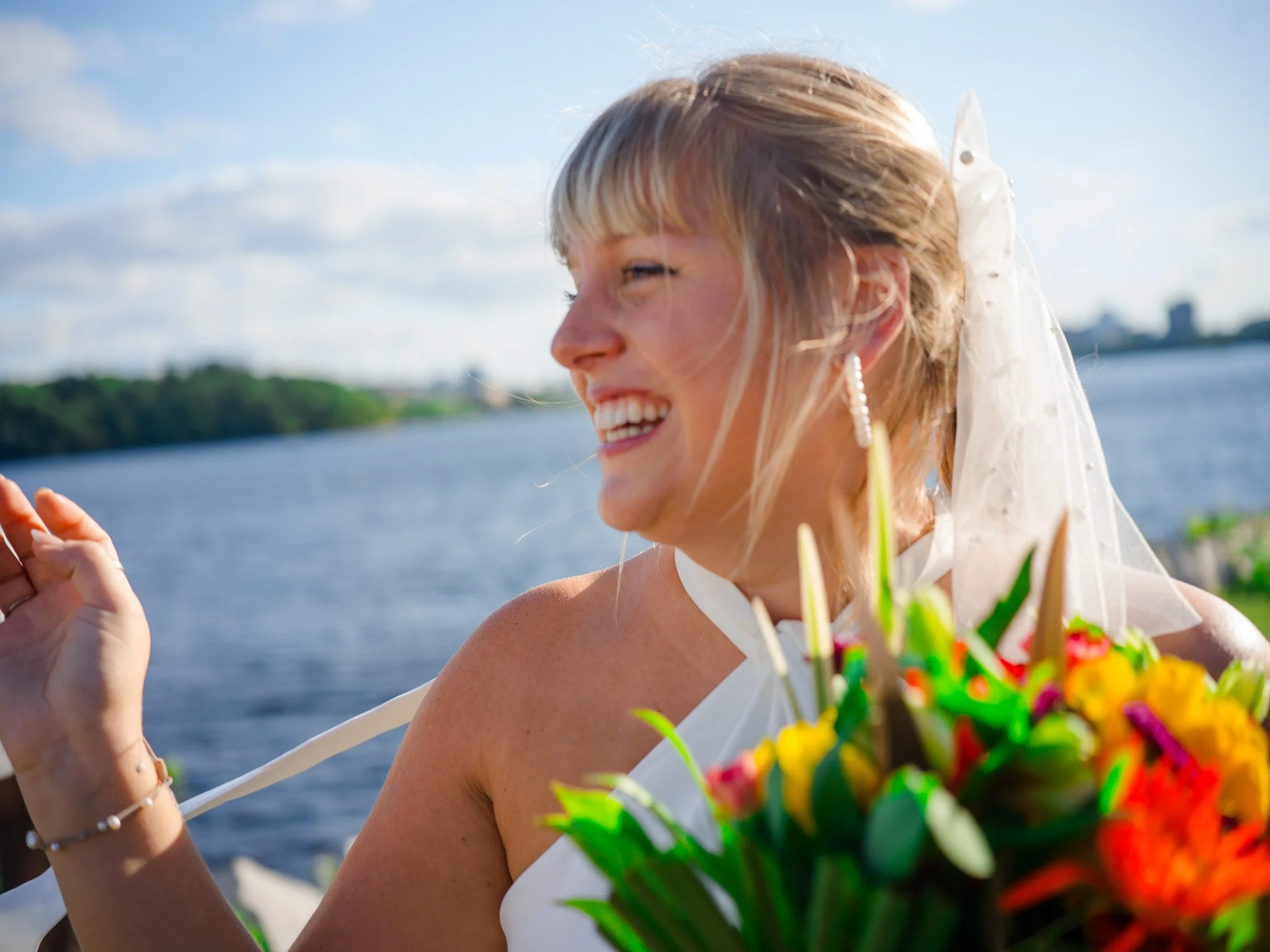 A woman in a wedding dress with a white veil smiling and holding a bouquet of colorful flowers near a body of water with trees in the background.