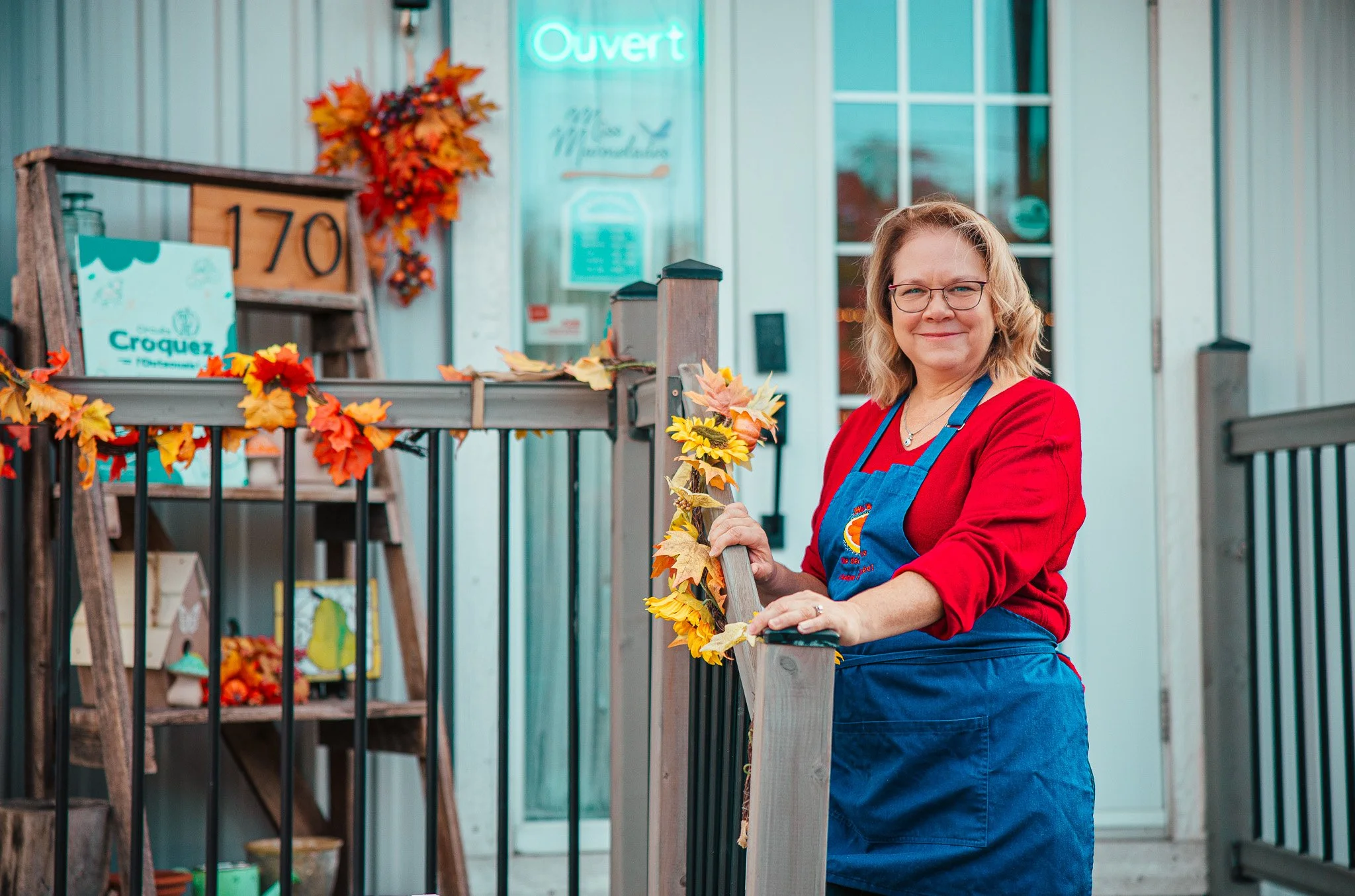 A woman in a red shirt and blue apron stands outside a shop, holding onto a wooden railing decorated with fall leaves and sunflowers, smiling at the camera.