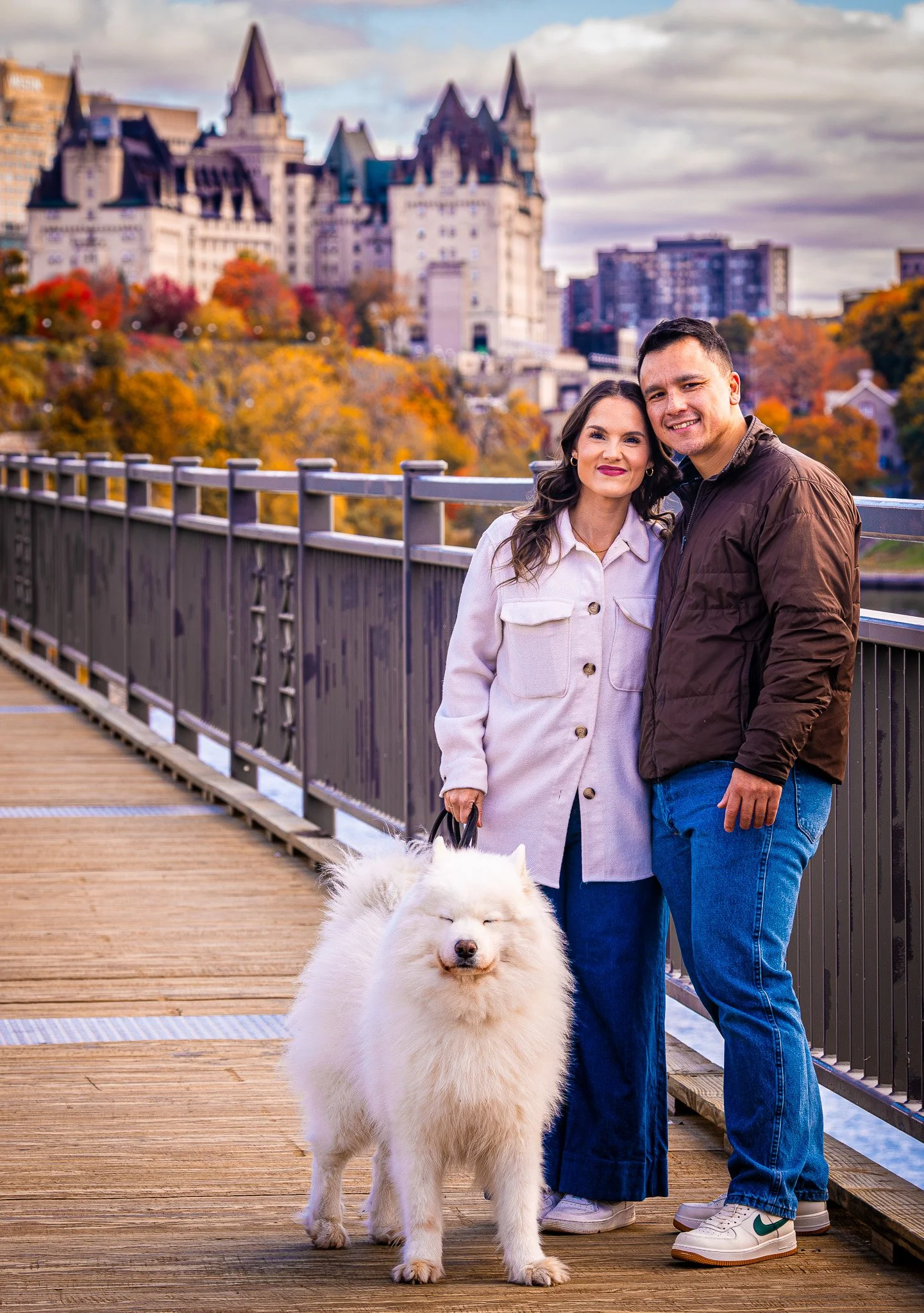 A couple standing on a wooden bridge with a white fluffy dog. They are smiling with autumn foliage and a castle-like building in the background.