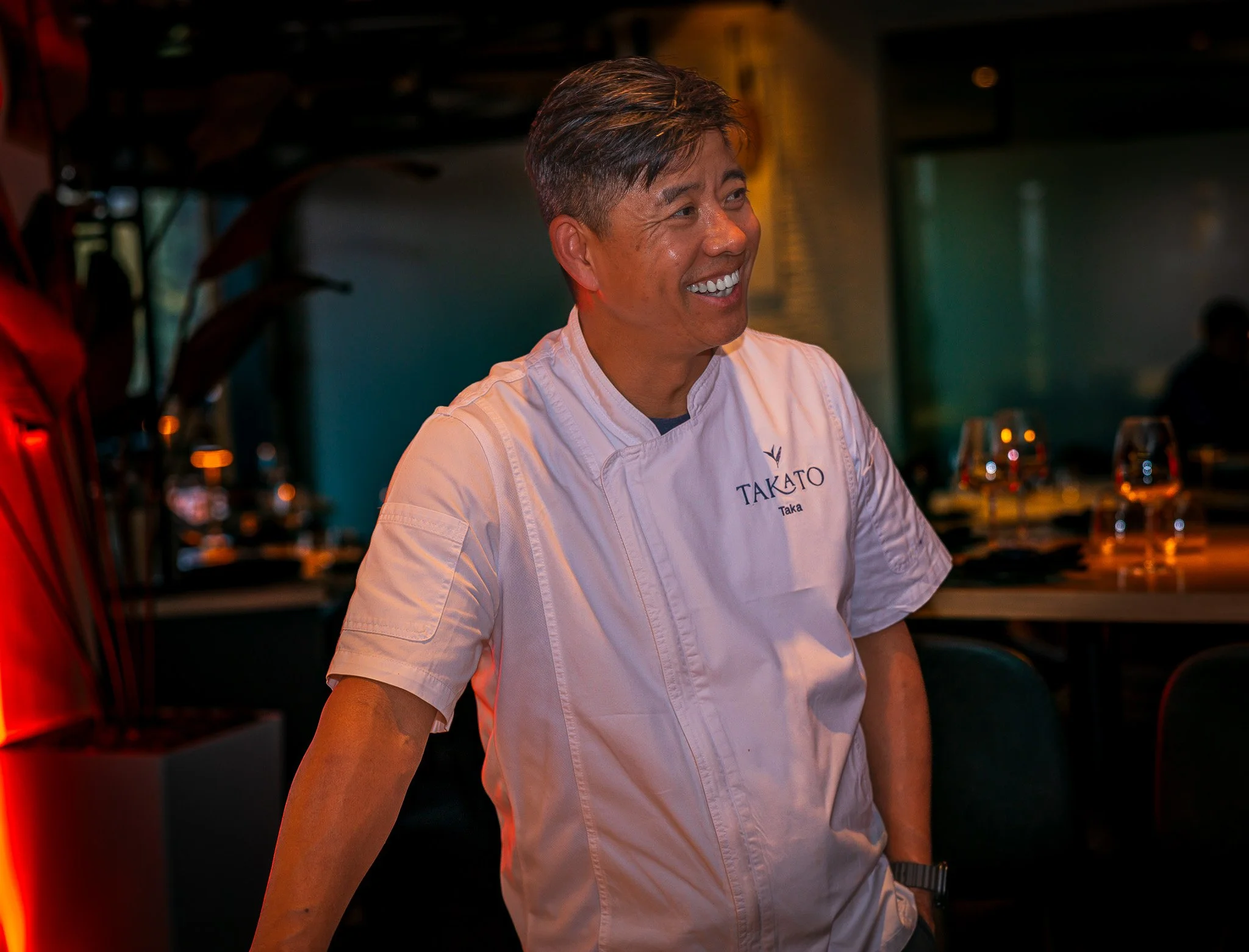Smiling male chef wearing a white coat with 'Takato' embroidered on it, in a restaurant setting with glasses on tables in the background.