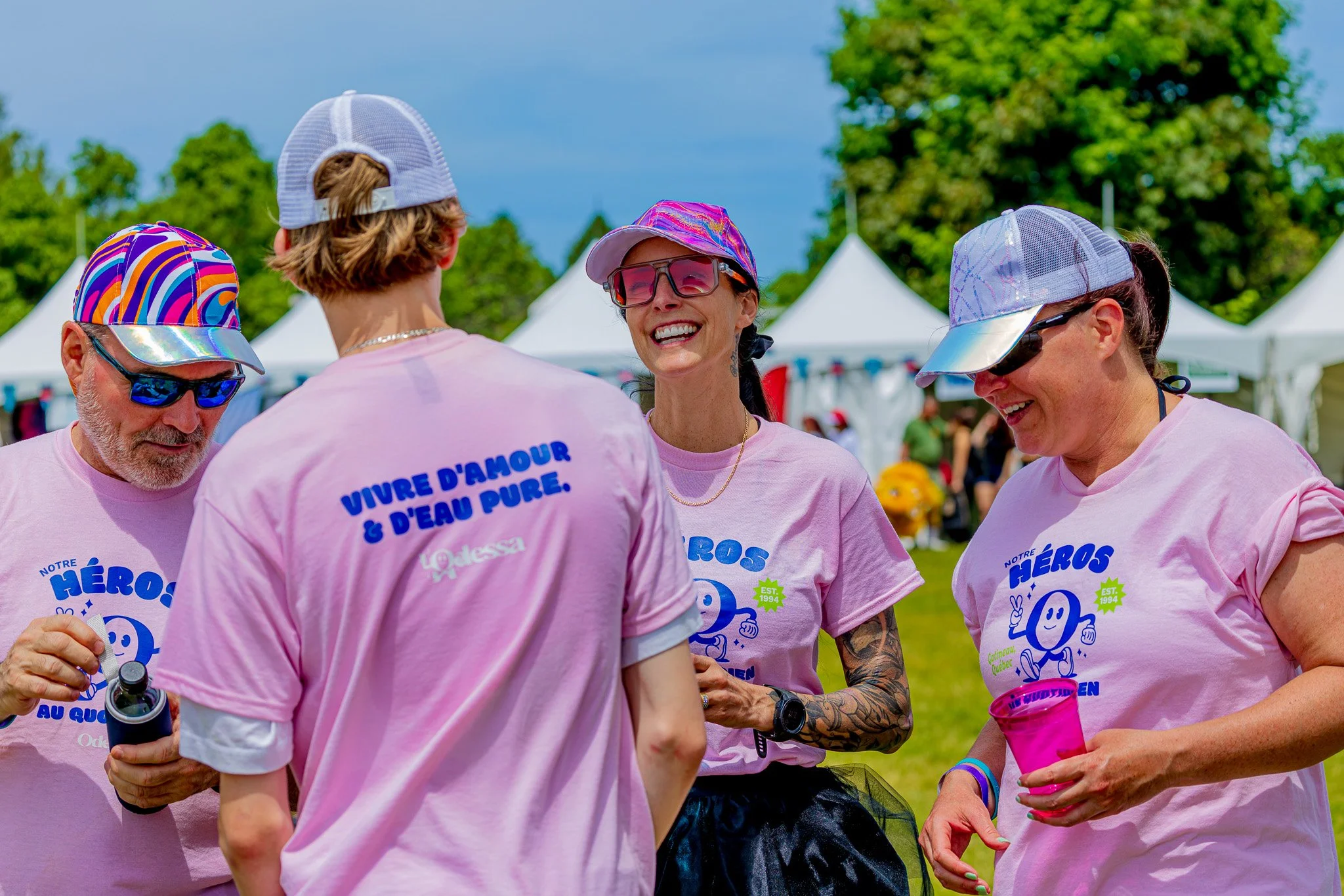 Group of four women and one man, all wearing pink shirts and colorful caps, gathered outdoors at a summer event with white tents in the background, smiling and chatting.
