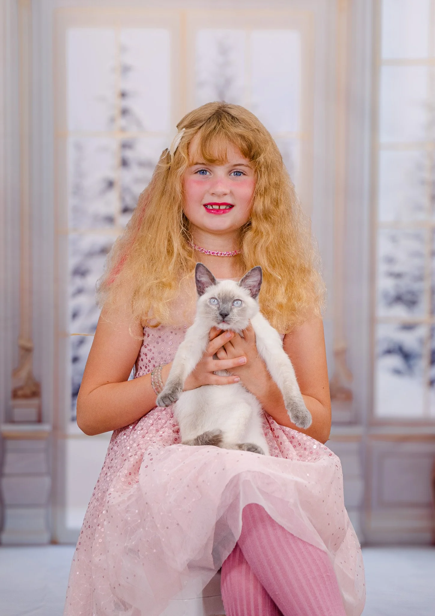 A young girl with long, curly red hair, wearing a pink dress and striped pink tights, is sitting and holding a Siamese cat with blue eyes in front of snowy windows.
