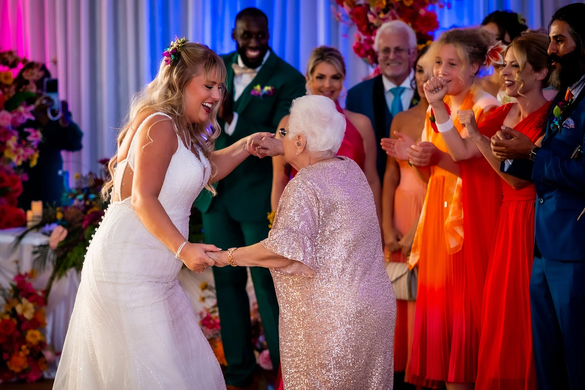 A bride holding hands with an elderly woman during a wedding celebration with guests clapping in the background.