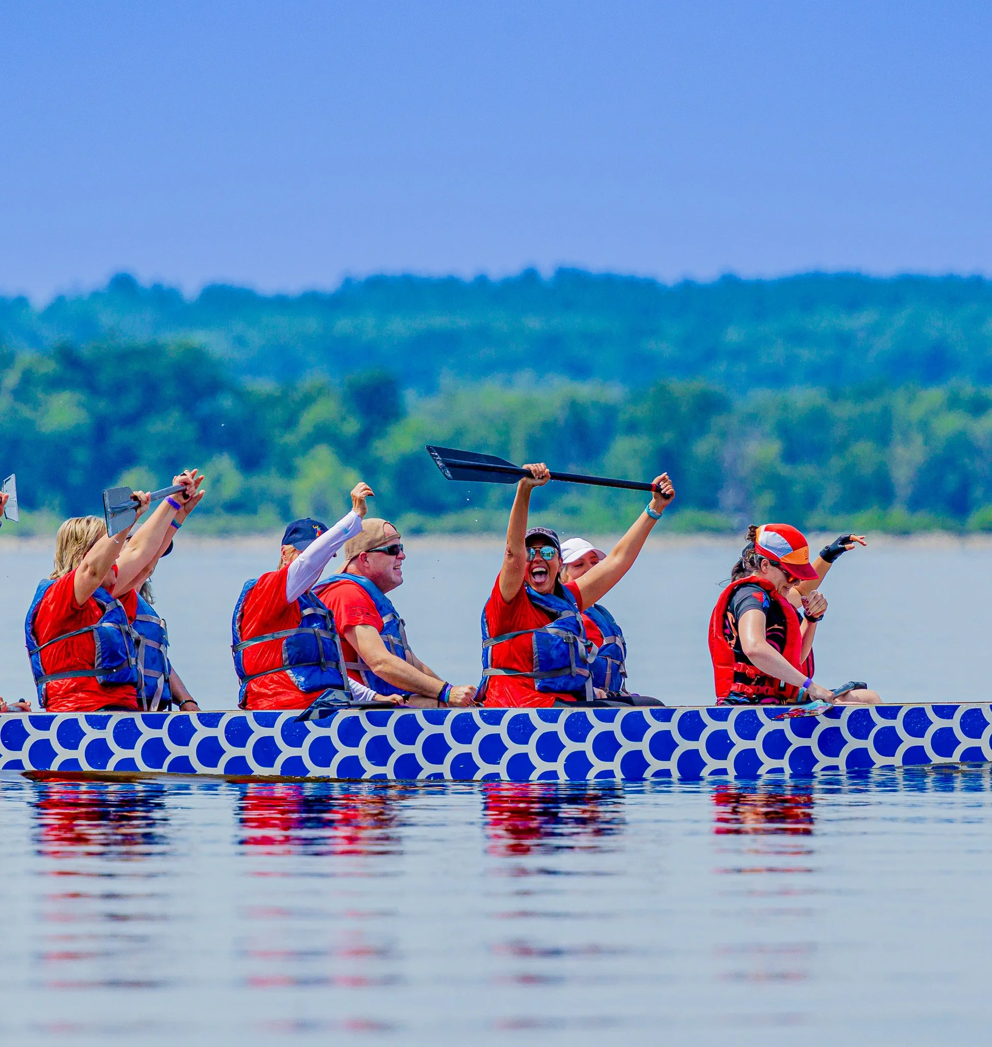 People in a dragon boat race on water, wearing red and blue life jackets, with some holding paddles and celebrating.