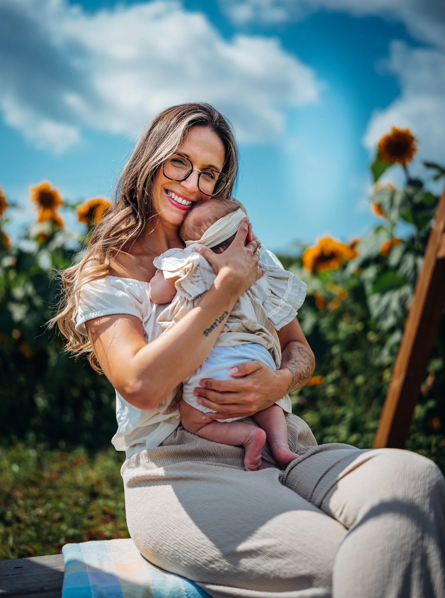 A woman with long hair, glasses, and tattoos happily holding a newborn baby outdoors among sunflowers on a bright, partly cloudy day.