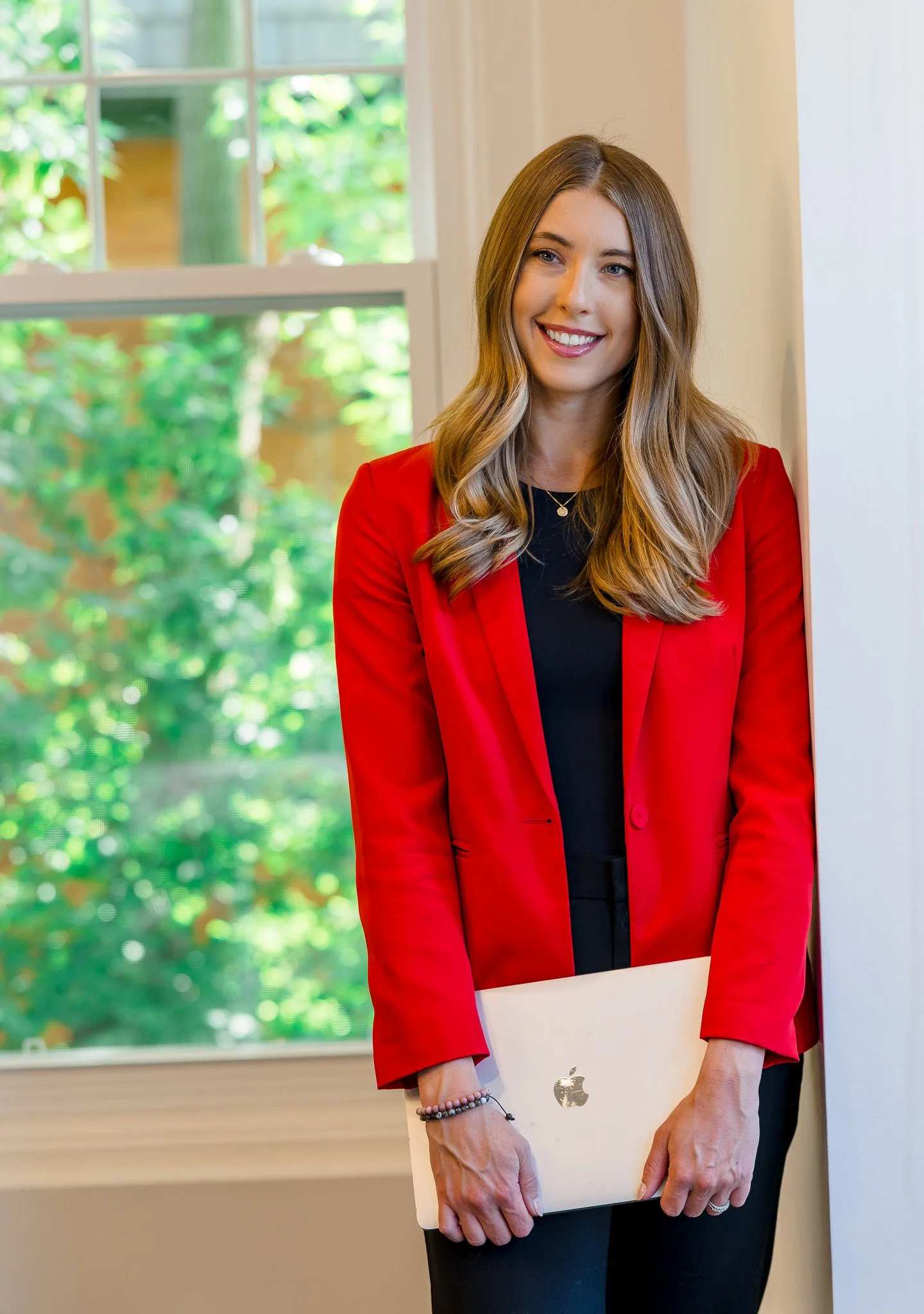 A woman with long light brown hair wearing a red blazer and black top holding a closed MacBook in front of her smiling, standing by a window with a green outdoor view.