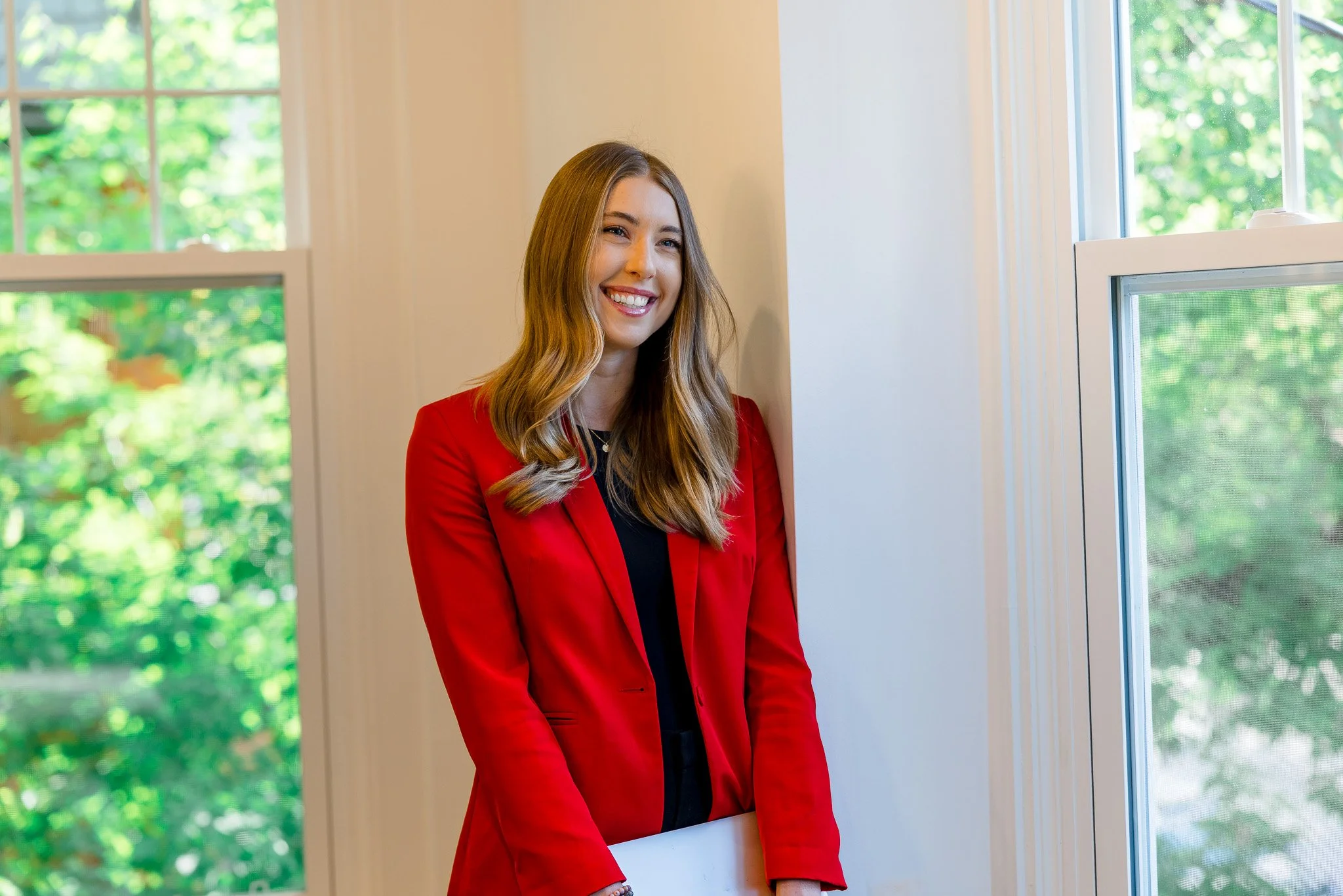 A woman with long light brown hair wearing a red blazer smiling while standing near a window with a view of green trees outside.
