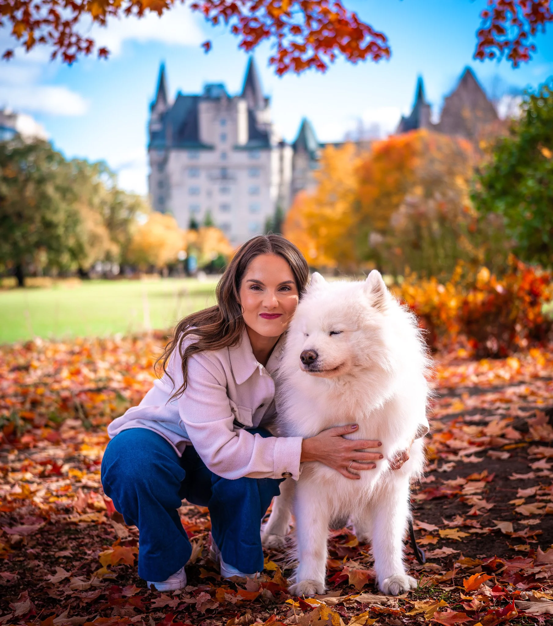A woman hugging a white fluffy dog in a park during autumn with colorful fall leaves scattered on the ground and a castle in the background.