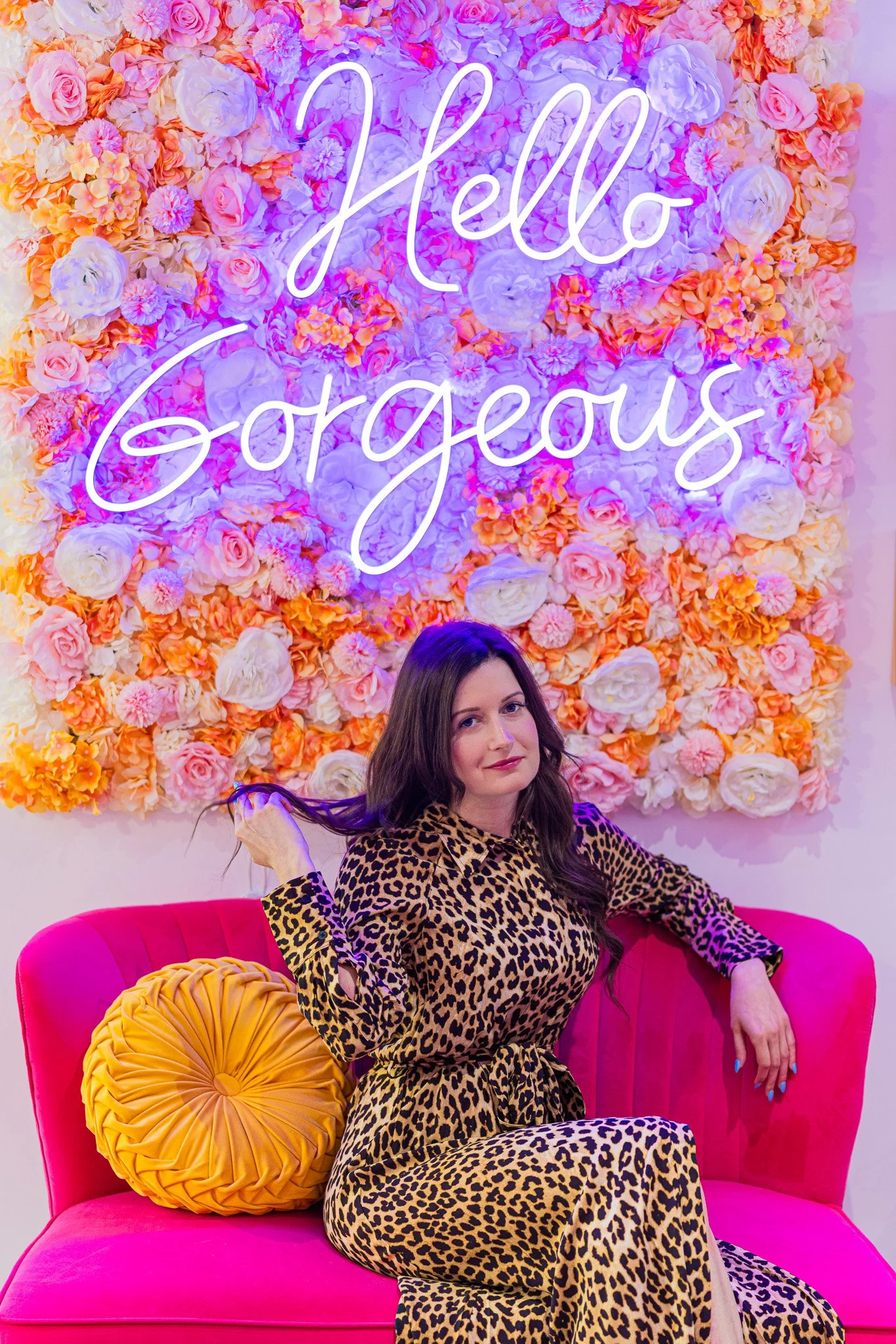 A woman sitting on a pink couch with a fluffy yellow pillow, wearing a leopard print dress, in front of a floral wall with a neon sign saying 'Hello Gorgeous'.
