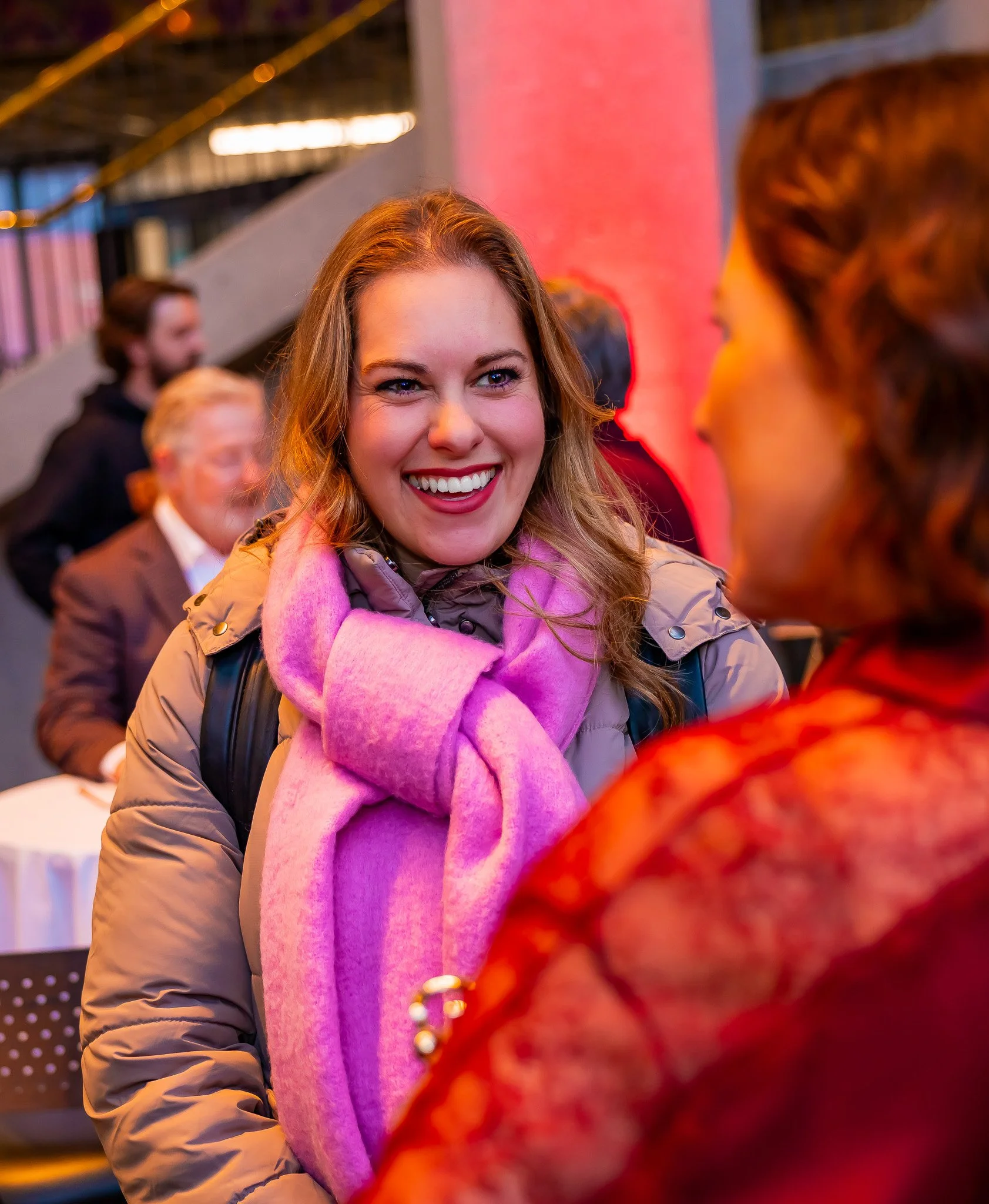 A smiling woman with wavy hair wearing a pink scarf and a tan jacket, engaging in conversation with another woman in an indoor social setting.