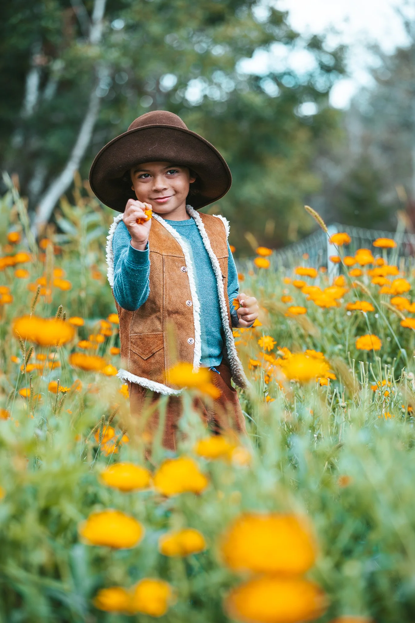 A young boy wearing a wide-brimmed hat and a brown vest standing in a field of orange flowers, holding one flower in his hand and smiling at the camera.