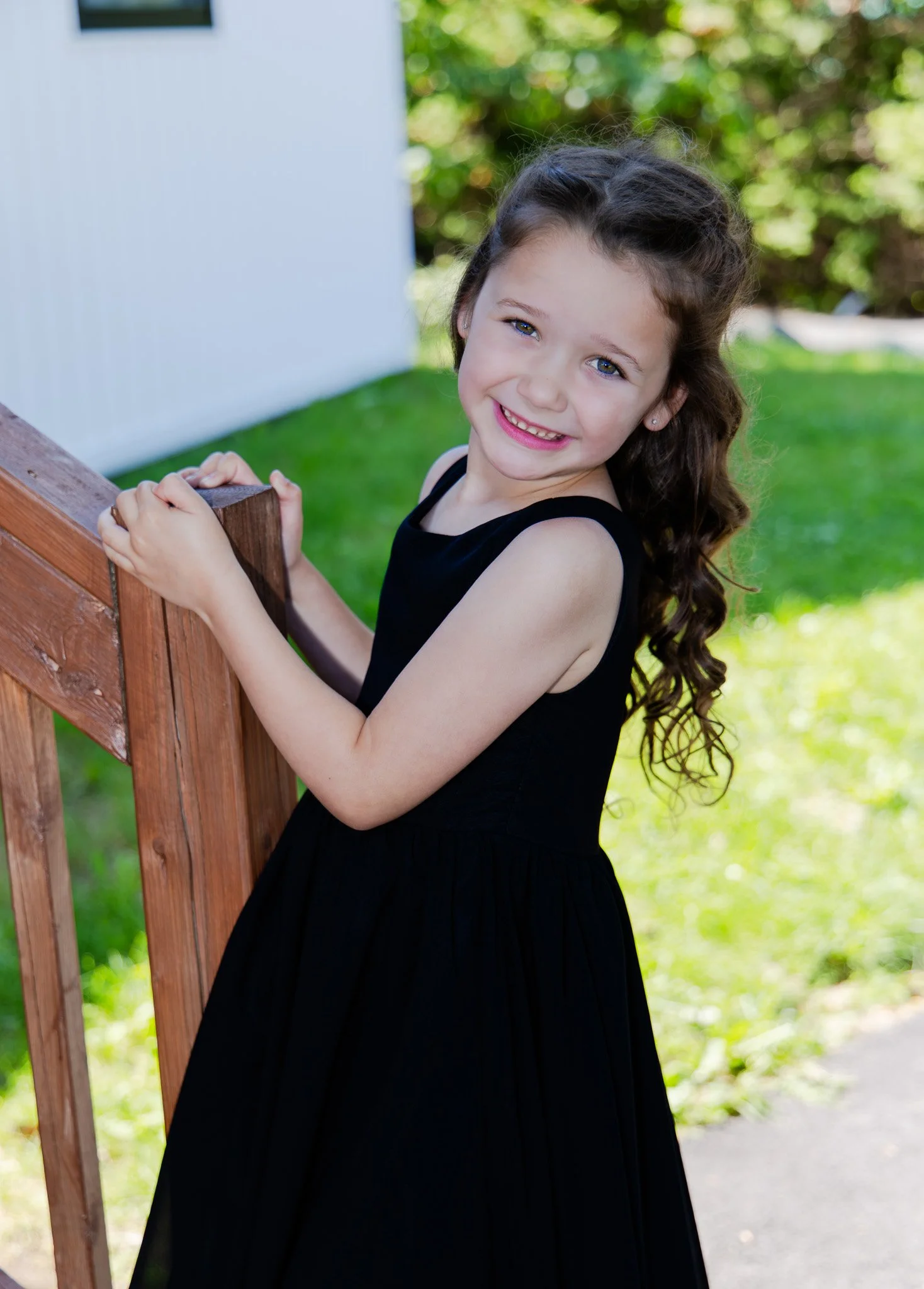 A young girl with curly brown hair and blue eyes smiling while holding onto a wooden railing outdoors, with green grass and trees in the background.