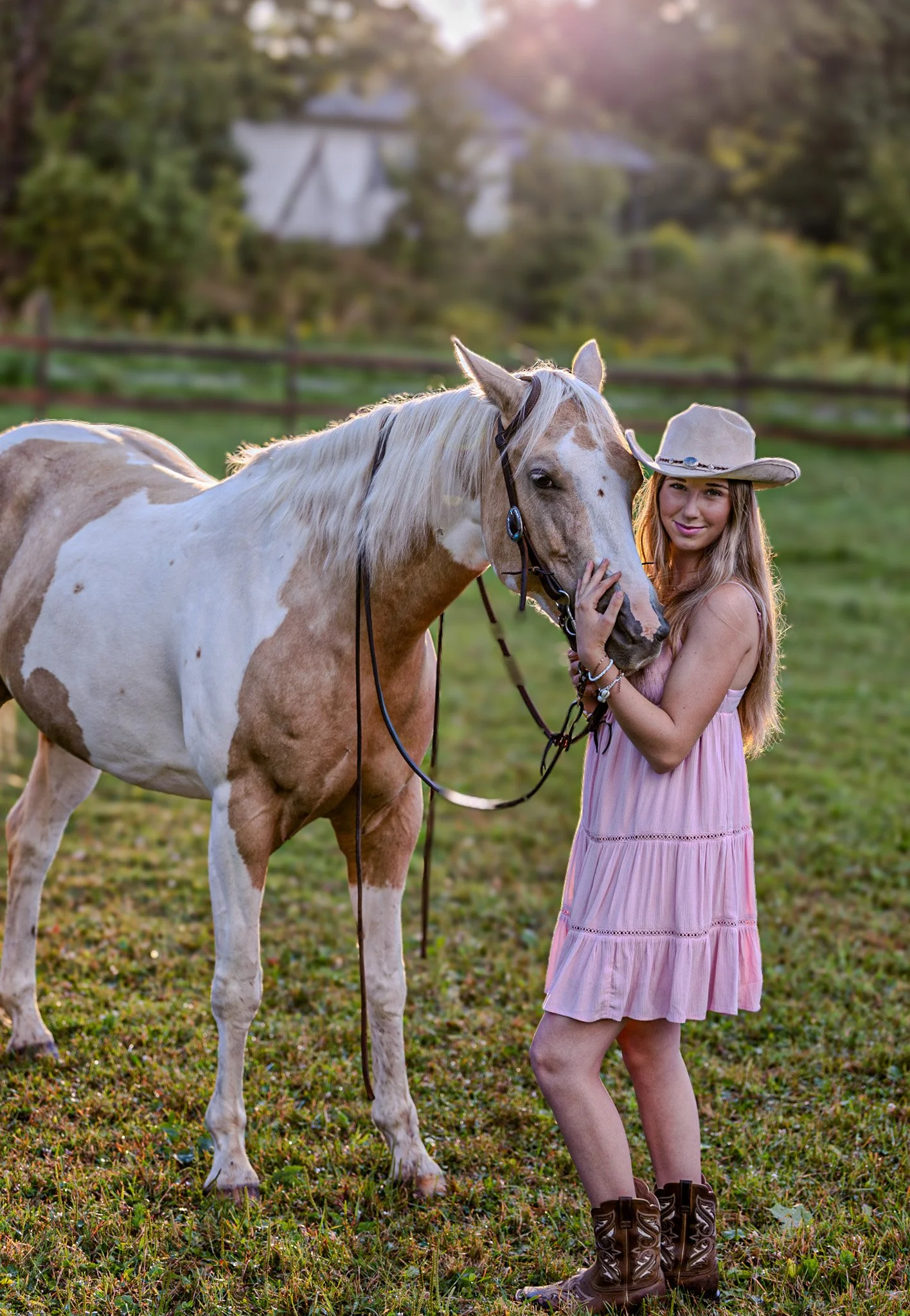 A woman in a pink dress and cowboy hat hugging a white and brown paint horse in a grassy field at sunset.