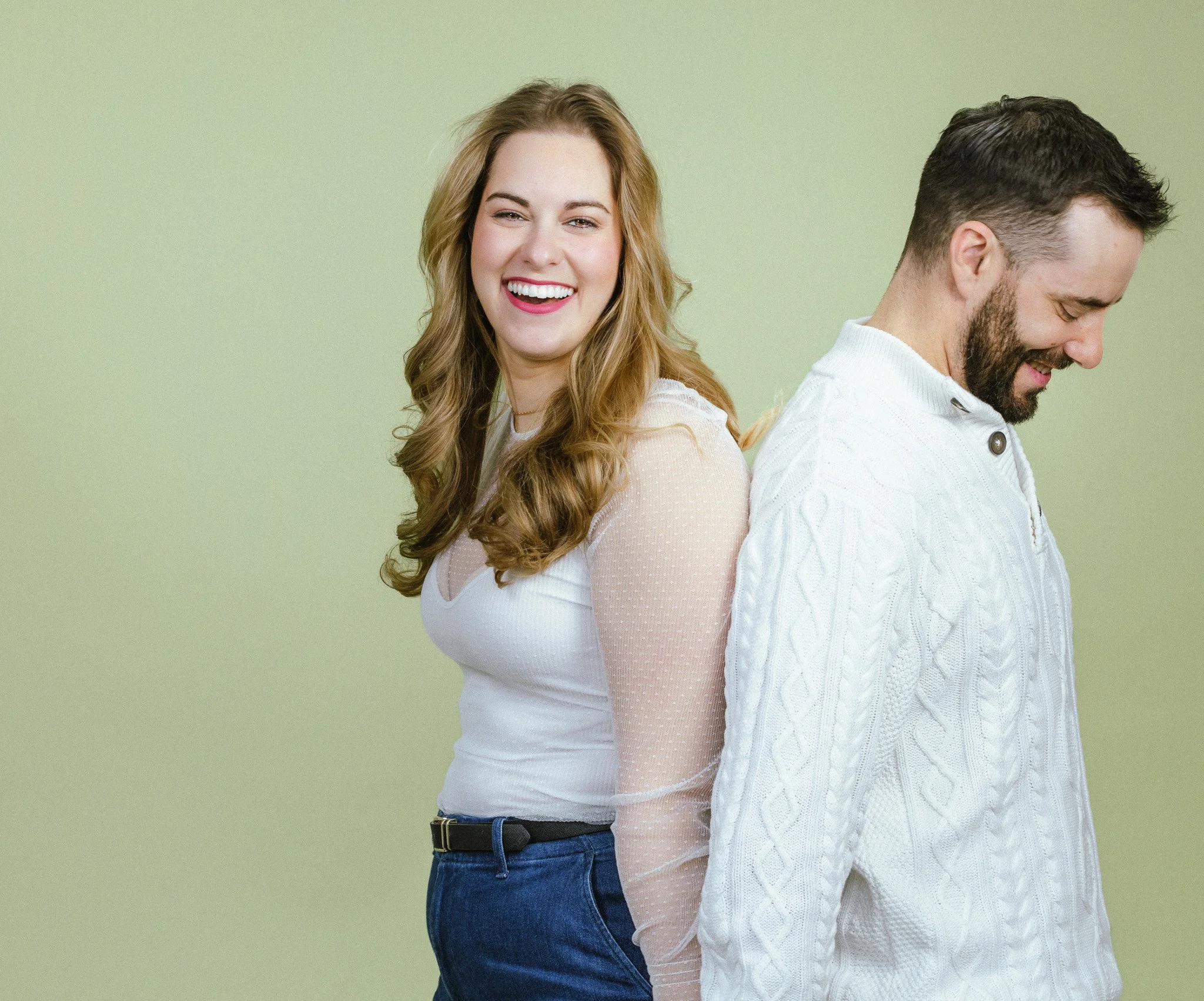 A smiling woman with long, wavy blonde hair stands behind a man with short dark hair and a beard. They are back to back, with the woman facing the camera and the man looking down.