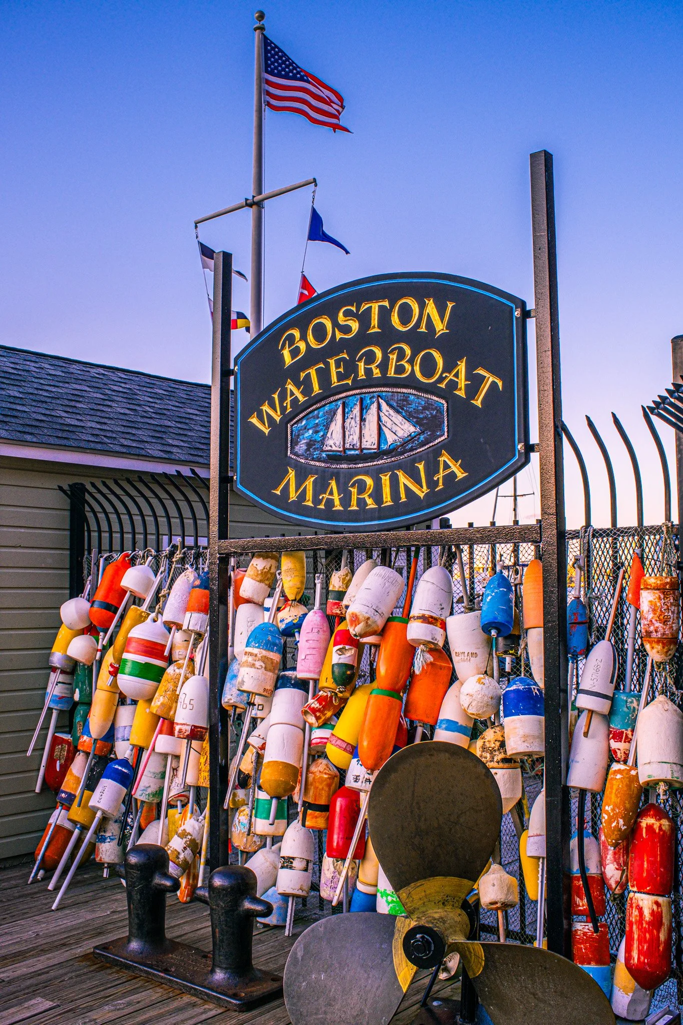 Sign at Boston Waterboat Marina with colorful buoys hanging on a fence and flags against a blue sky.