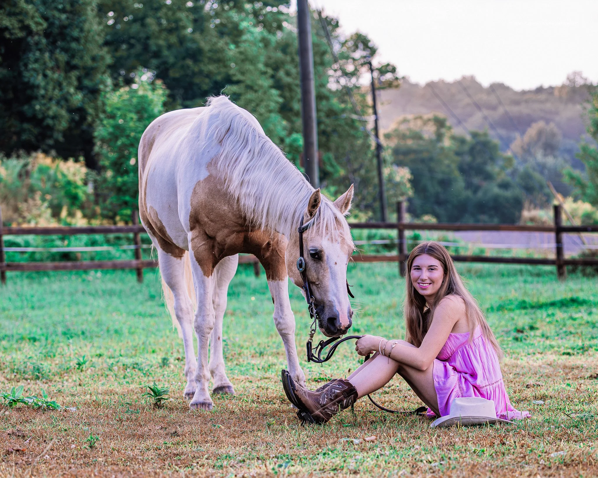 A young woman in a pink dress and brown boots sitting on the grass, holding the reins of a white and brown pinto horse in a fenced pasture with trees and hills in the background.