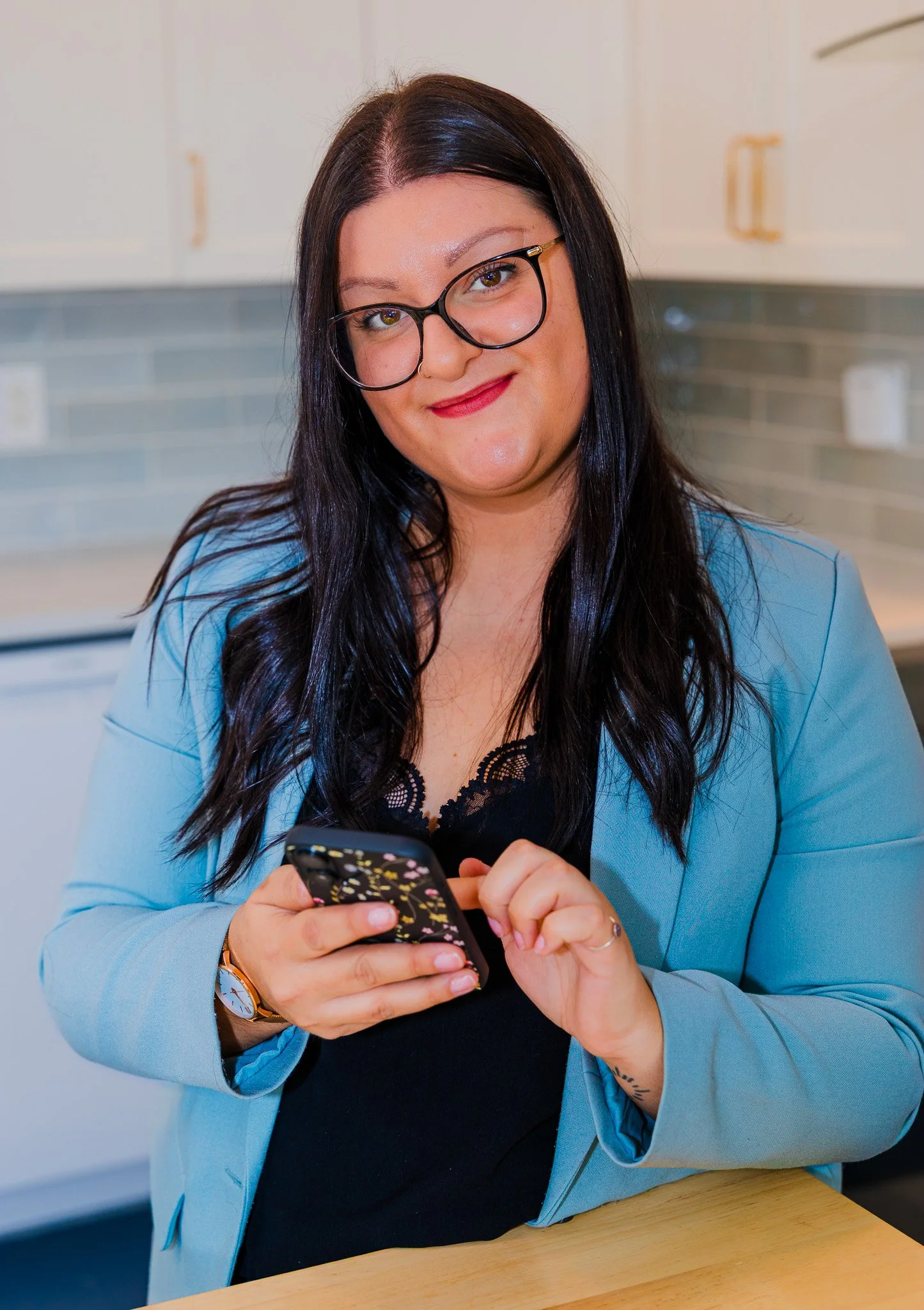 A woman with long dark hair, glasses, and a blue blazer sitting at a kitchen counter, looking at her phone and smiling.