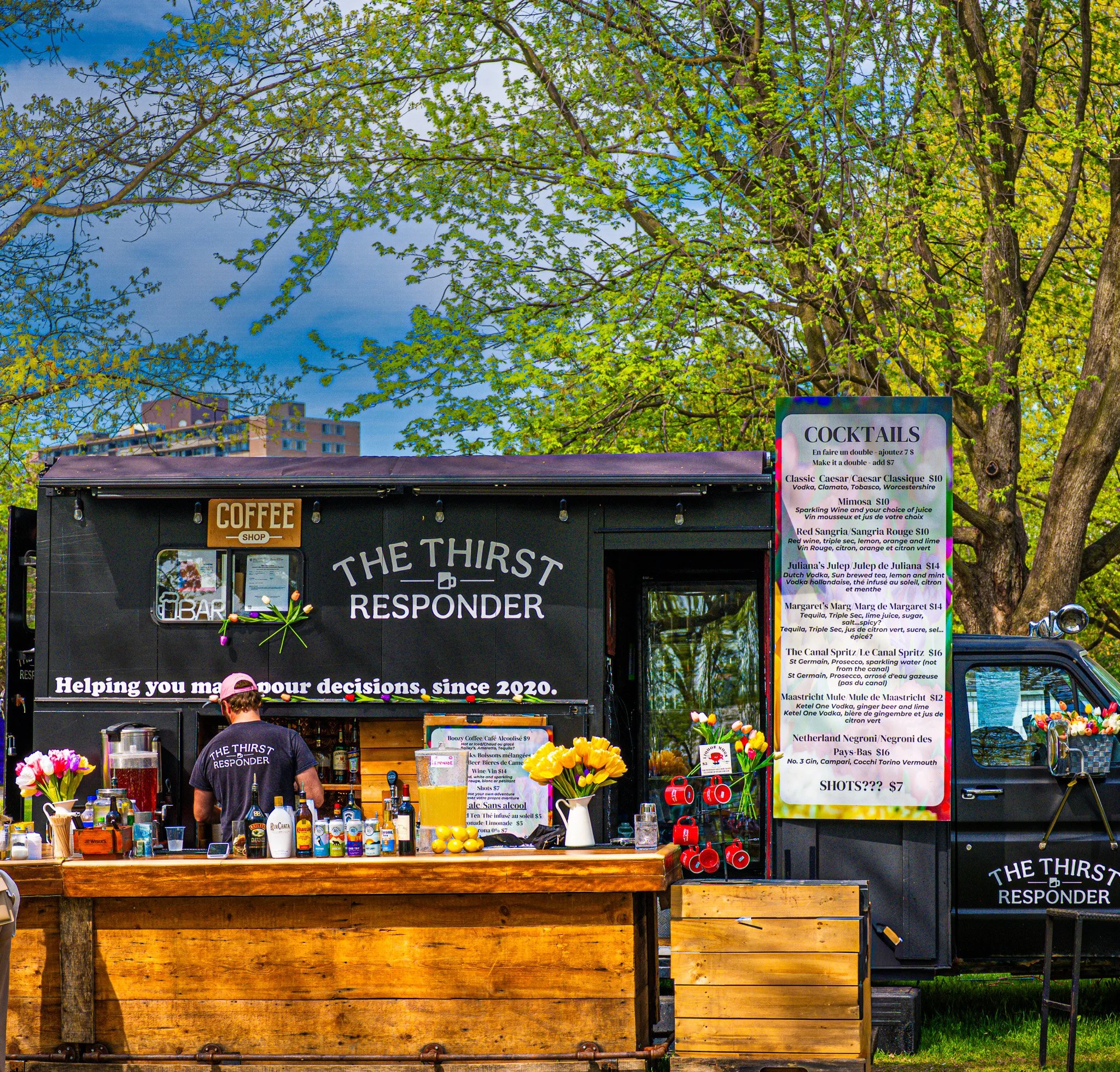 A black mobile coffee and cocktail bar with the sign 'The Thirst Responder' serves drinks at an outdoor location with green trees and a partly cloudy sky in the background. The counter has various bottles, a yellow drink, and flower arrangements, wit