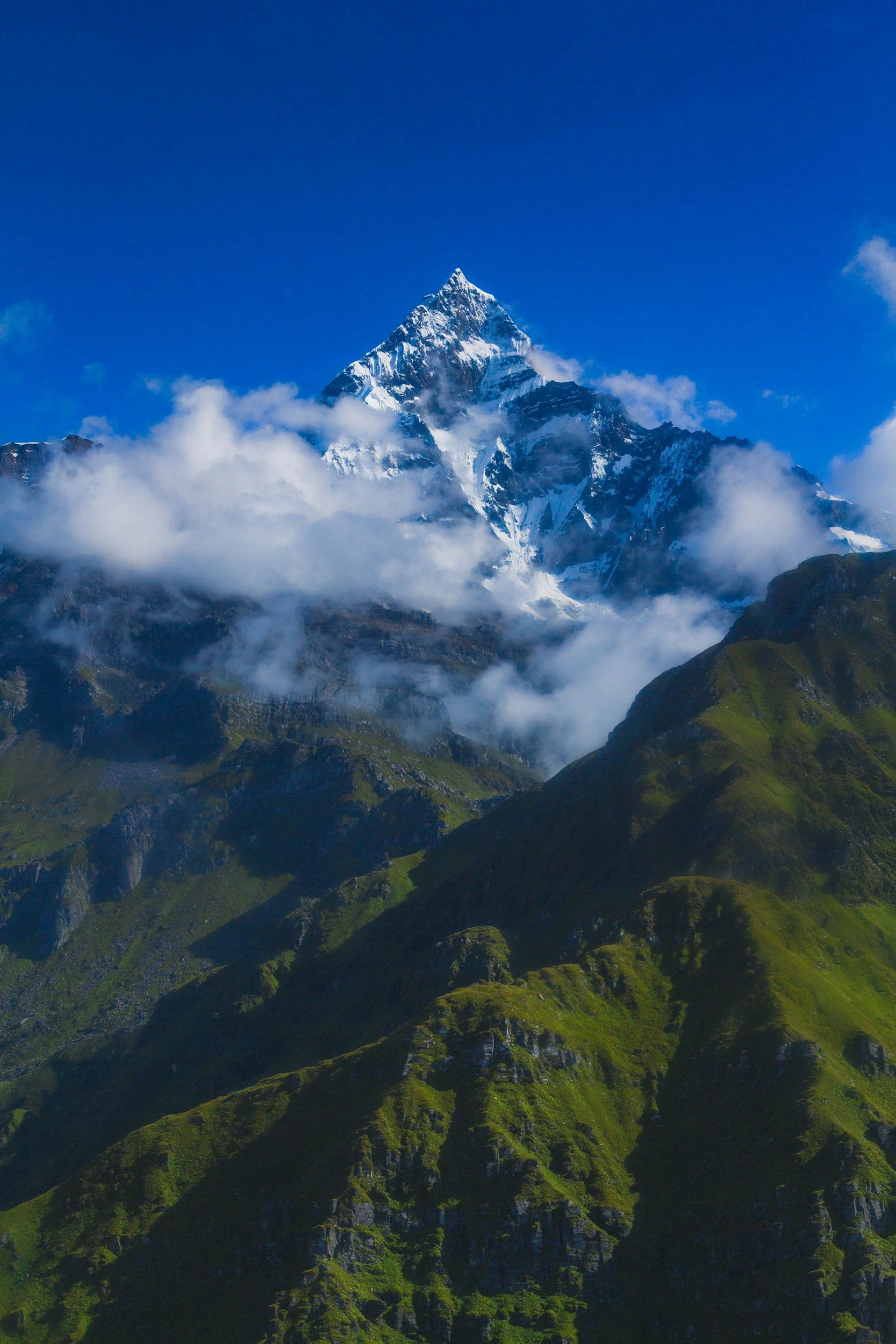 Snow-capped mountain peak with clouds and green hills in the foreground.