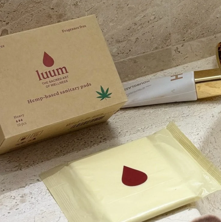 A box of hemp-based sanitary pads with the brand name 'Luum' on it, a green cannabis leaf icon, and the note 'fragrance free', placed on a bathroom countertop. A silver and gold razors are visible in the background.