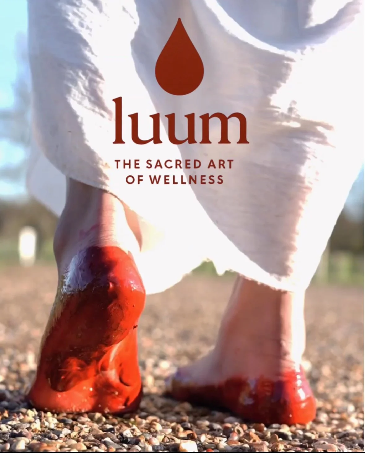 Close-up of a person wearing white clothing and orange-red shoes standing on gravel with a blurred outdoor background. Text overlay reads 'luum: The Sacred Art of Wellness' with a red droplet icon above the word 'luum'.