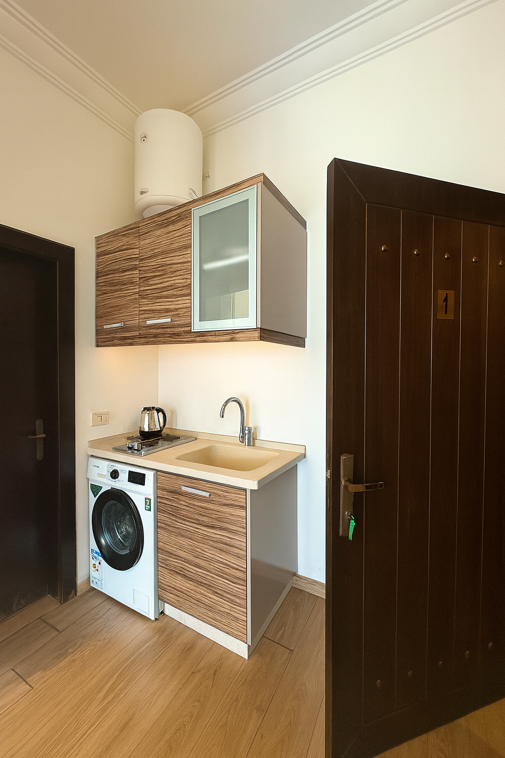 Small kitchenette area with a washing machine, a beige countertop with a sink, a small electric stove with a kettle, upper cabinets with wood finish and frosted glass door, a large white water heater on the ceiling, dark door with a lock, and wooden
