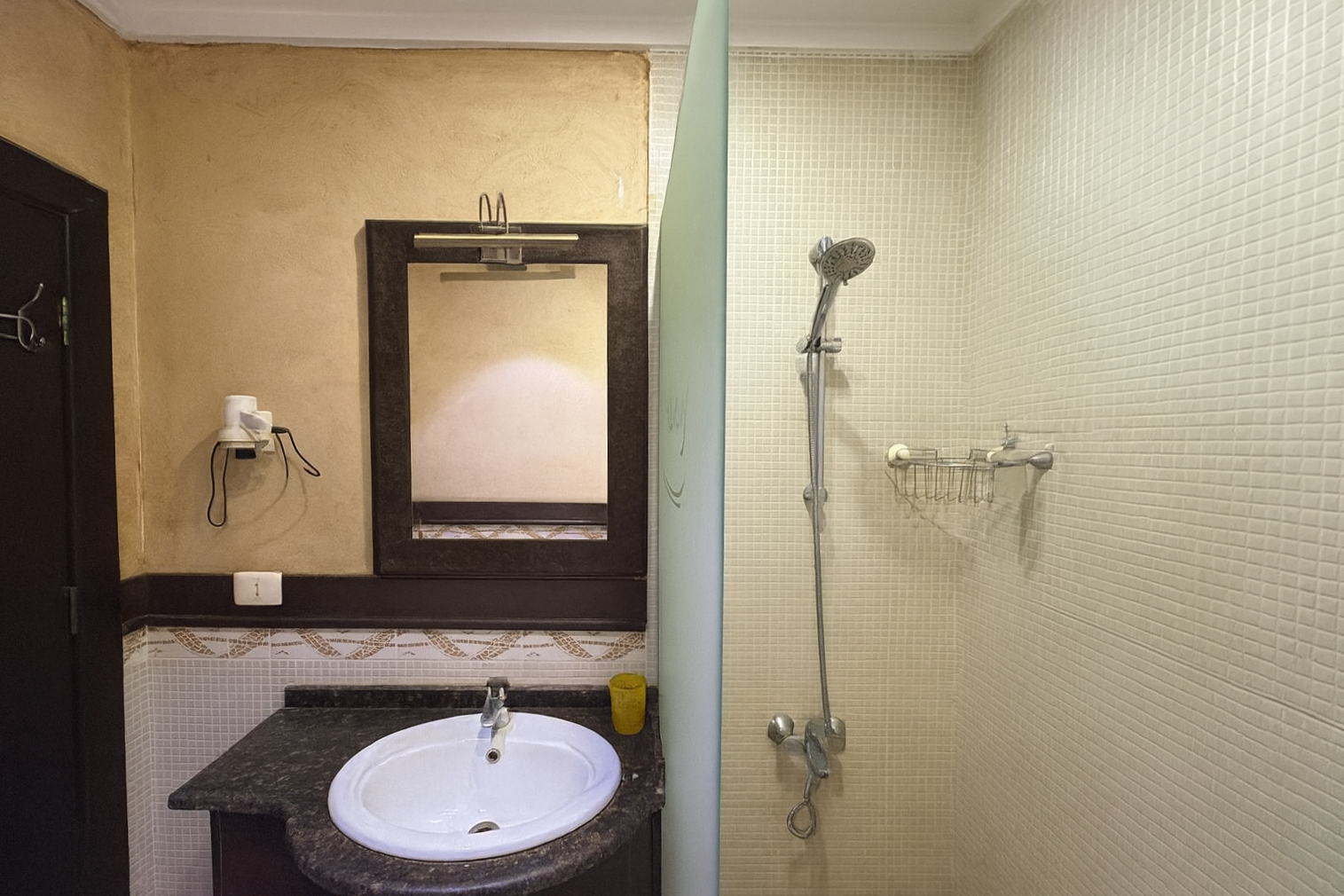 Bathroom split into two parts: on the left, a sink with a mirror and yellow wall; on the right, a shower area with tiled wall and a showerhead.