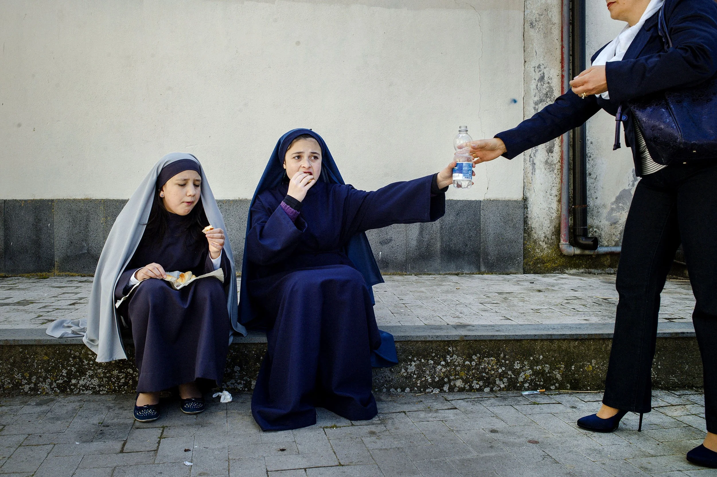 A mothers in high heels hands out water to her daughter during a break of one of the many processions during Easter. Randazzo, Sicily.