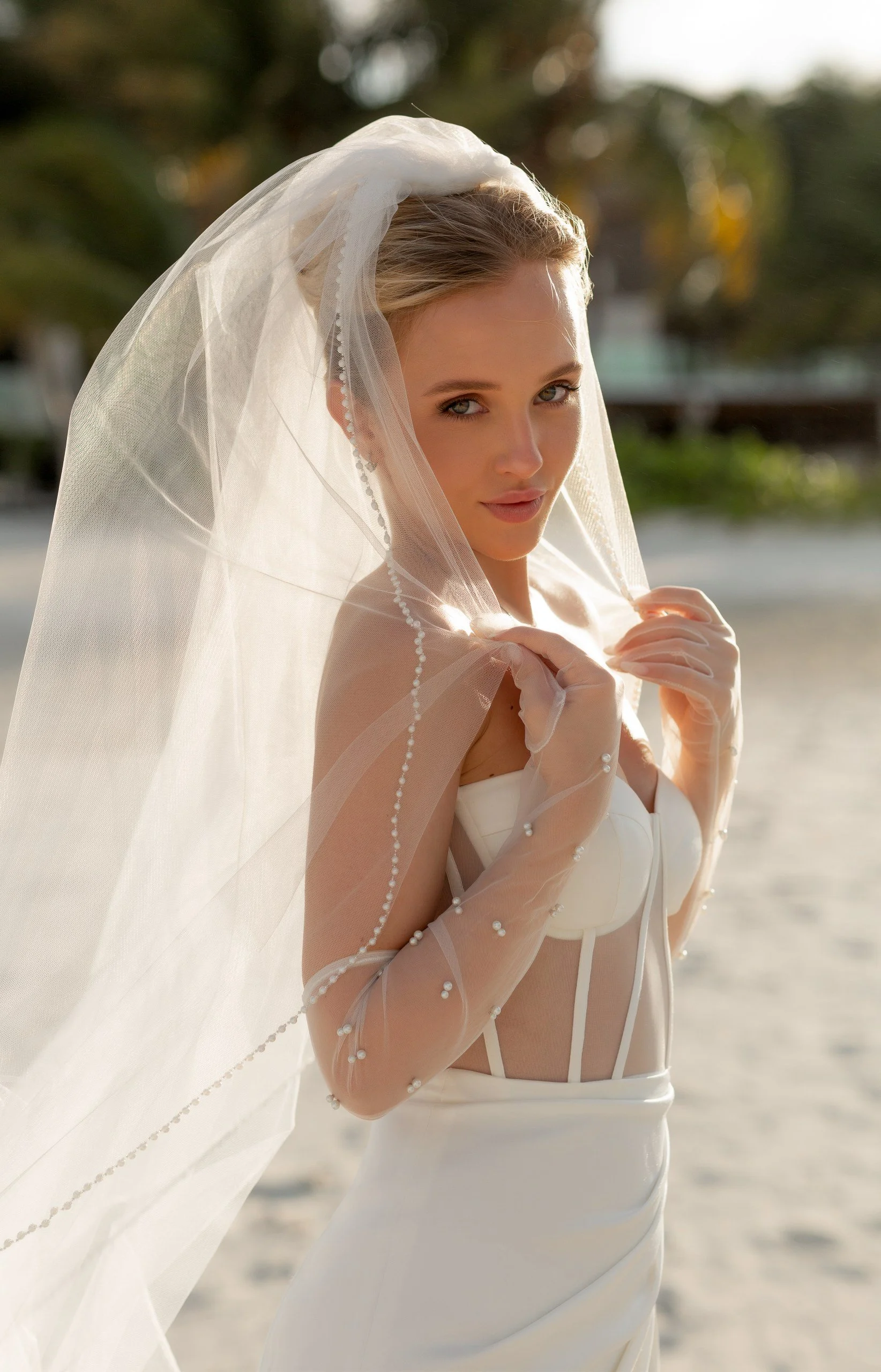 A woman in a wedding dress, holding a veil, outdoors near a body of water during sunset.