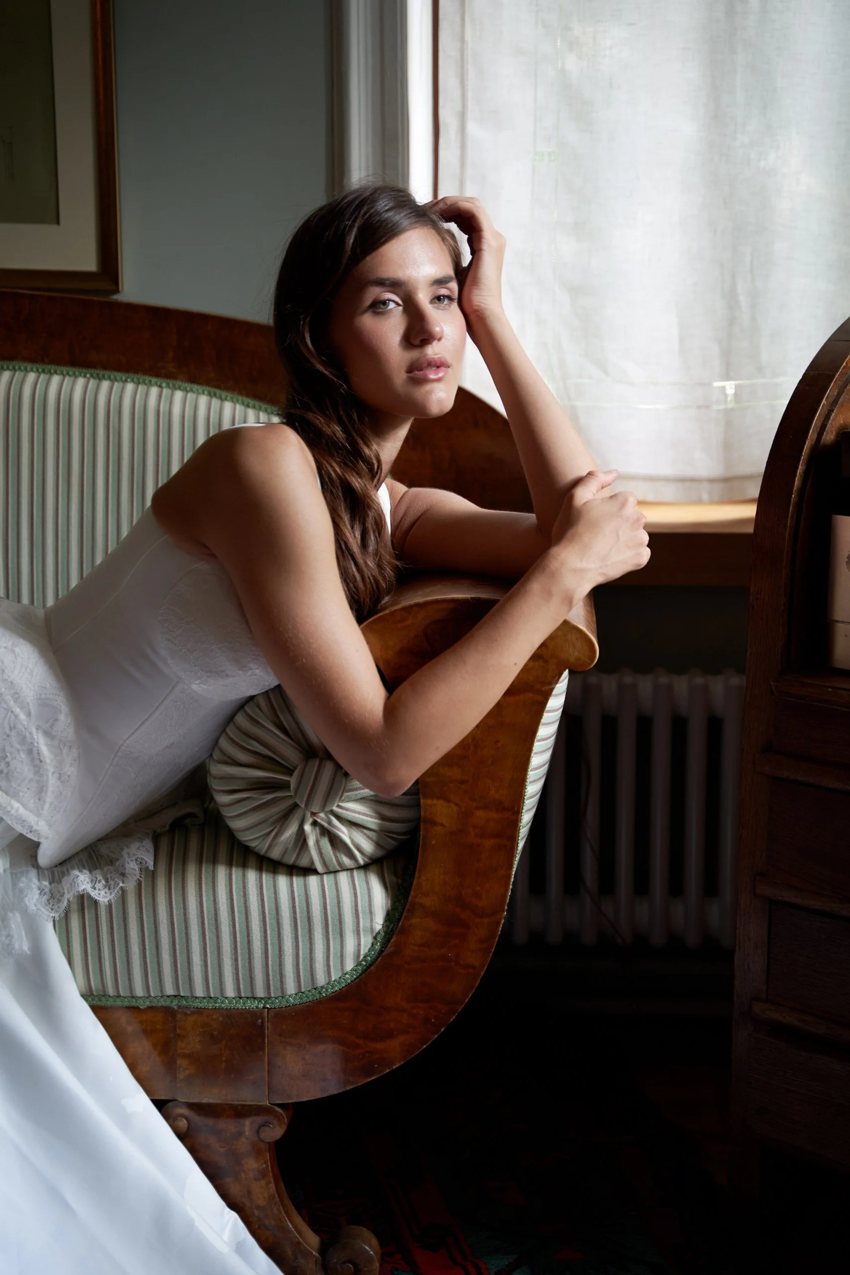 Young woman in a white dress lounging on a vintage wooden and striped cushioned sofa near a window, with her hand resting on her head and looking into the distance.