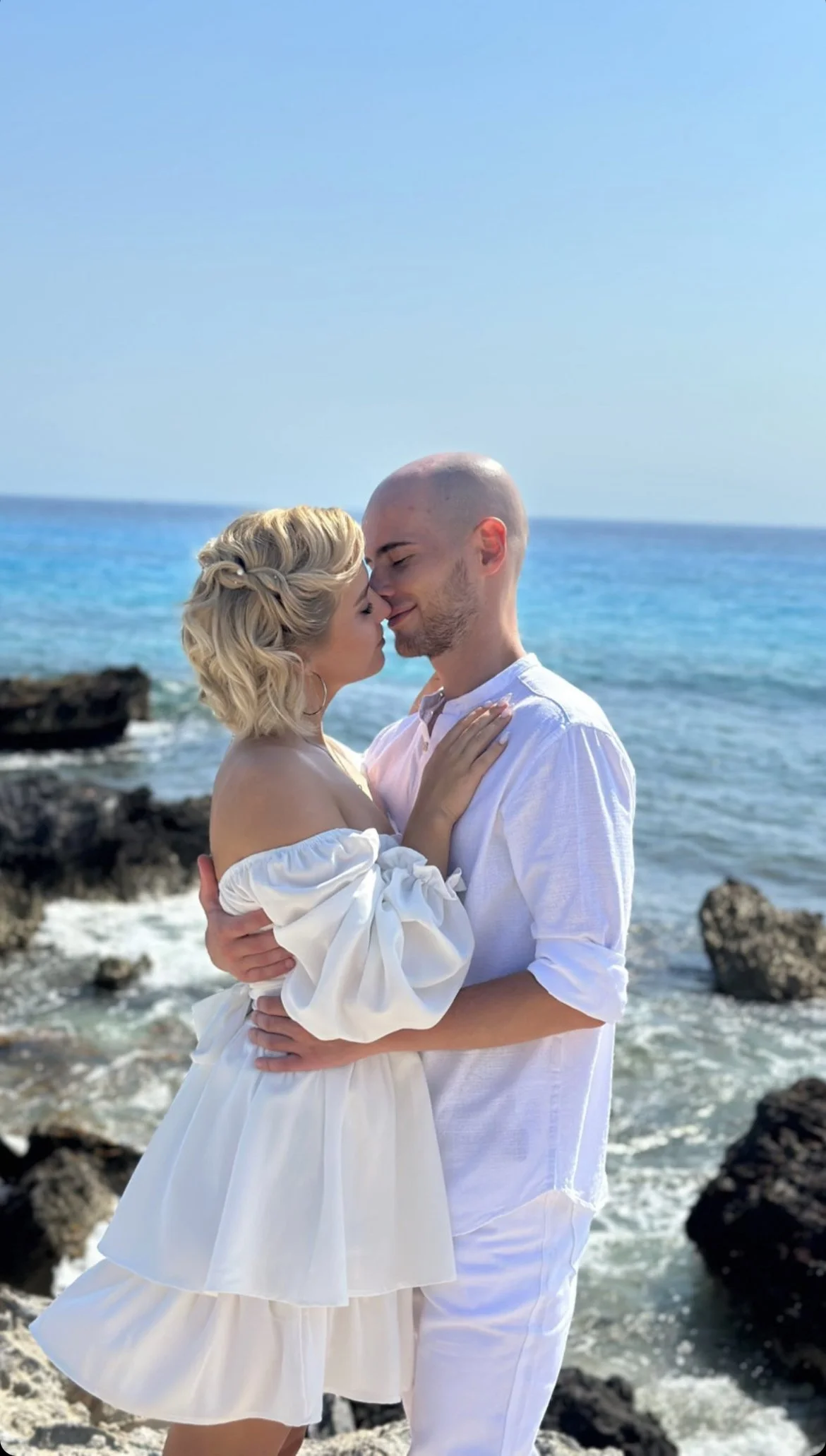 A couple standing on rocks by the ocean, embracing and touching foreheads, with the woman wearing a white off-the-shoulder dress and the man in a white shirt and pants, during daytime.