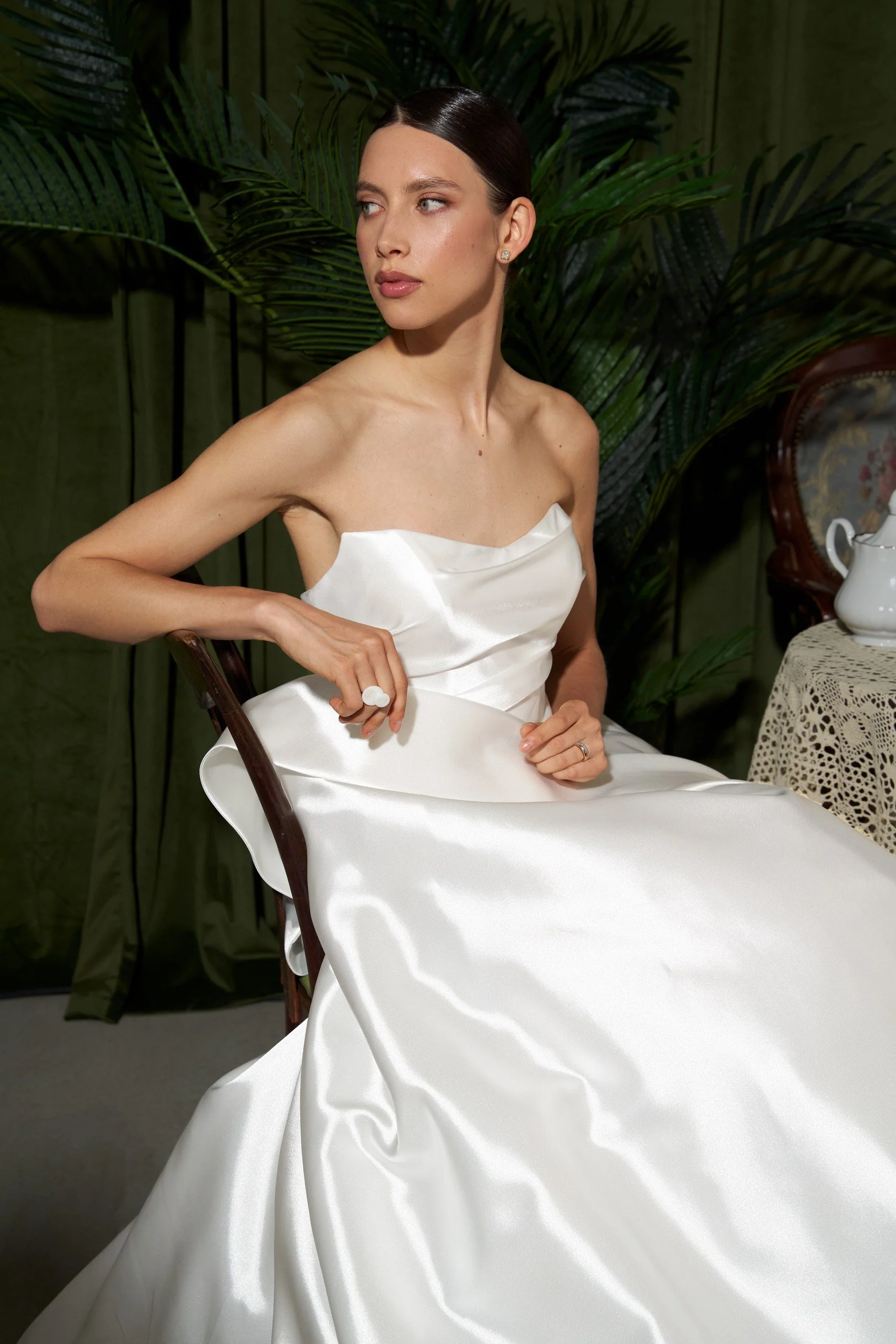 A woman in a strapless white satin gown sitting on a wooden chair in a room with green plants and a lace-covered table with a teapot.