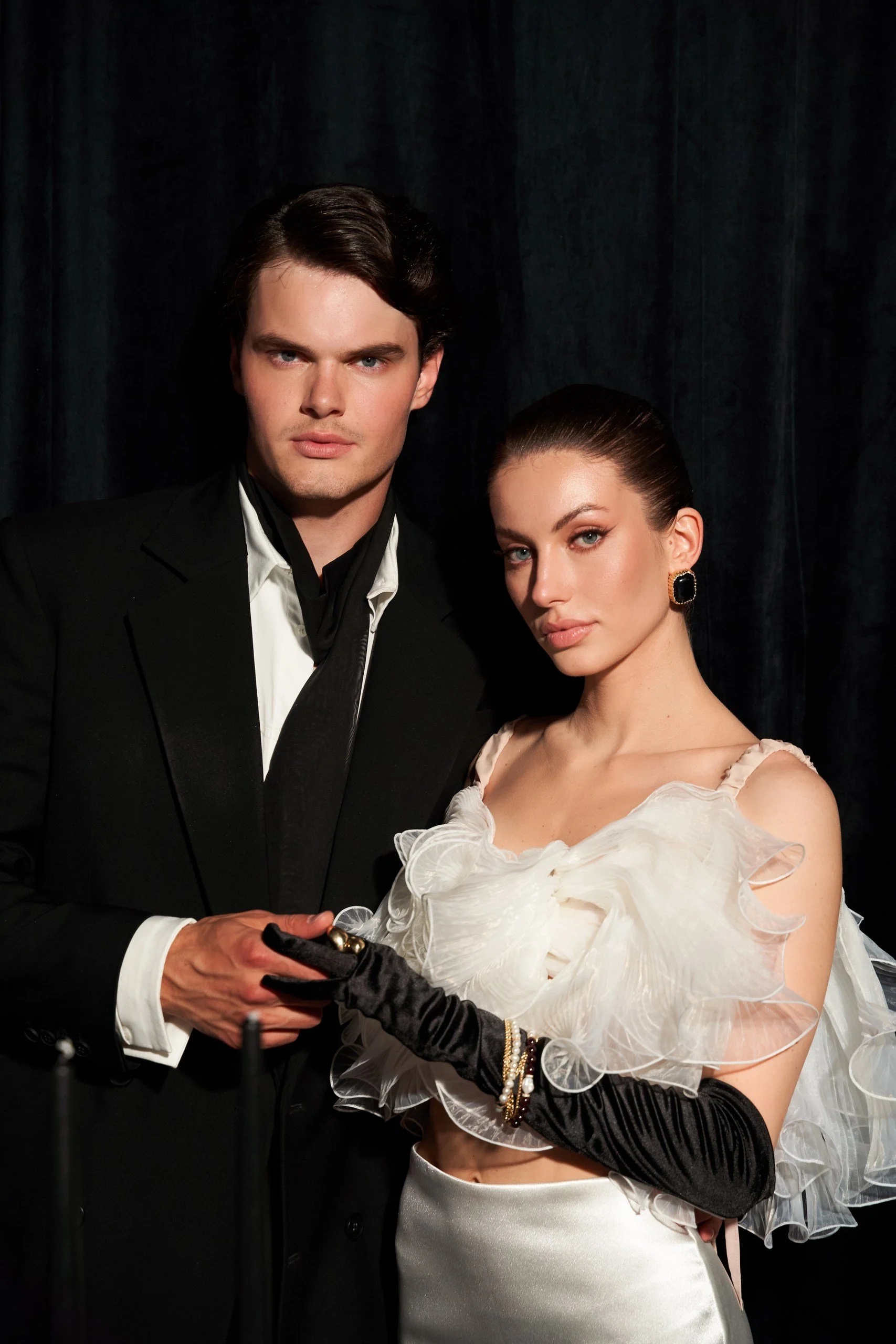 A man and woman dressed in elegant formal attire, holding hands, with dark background.
