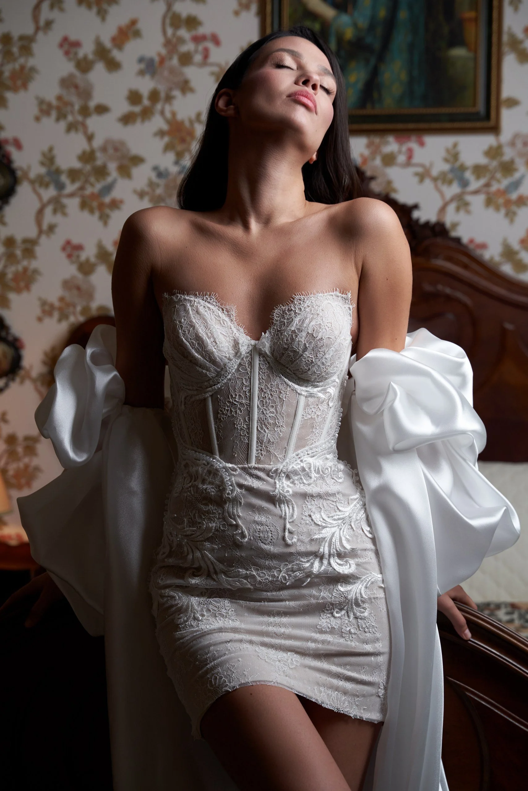 A woman wearing a white, lace, corset-style dress with puffed sleeves, sitting on a bed in a vintage bedroom with floral wallpaper.