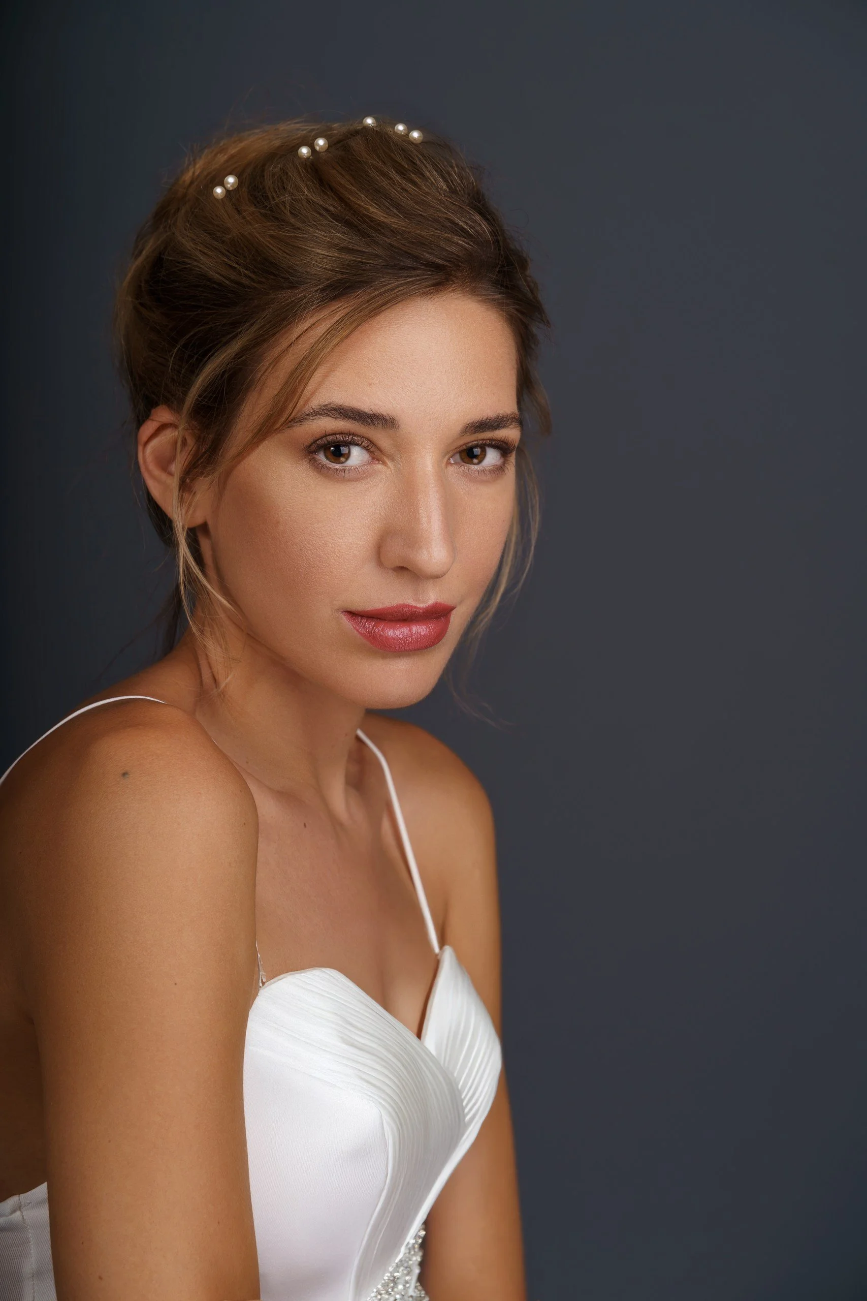 A young woman with brown hair styled upward, adorned with small pearls, and wearing a white dress with thin straps, posing against a dark background.