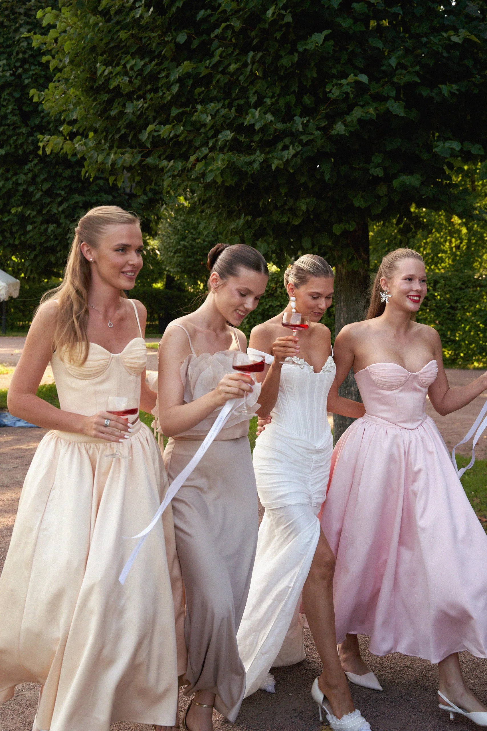 Four women in elegant dresses walking outdoors with glasses of red wine, surrounded by green trees and natural scenery.