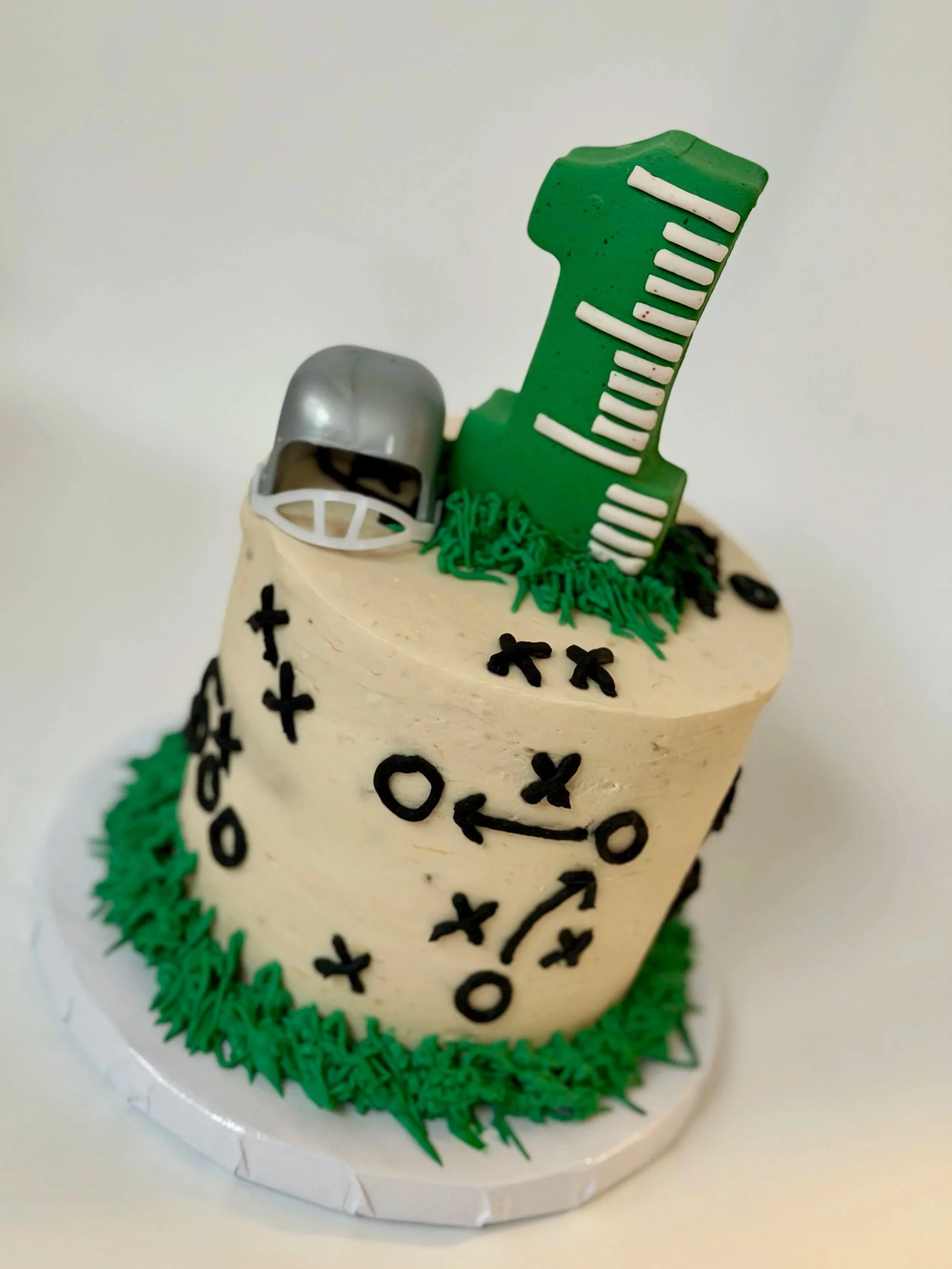 A sports-themed birthday cake decorated with football plays, with a large green number one as a topper.