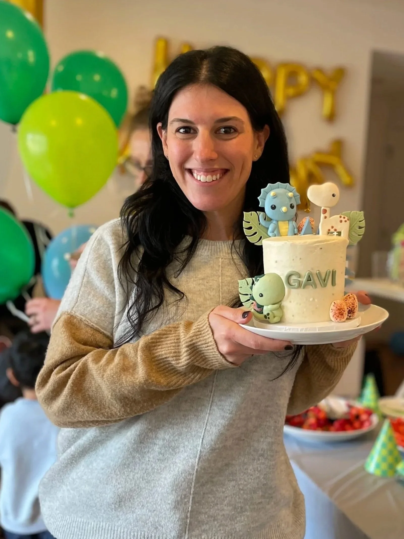 Woman with dark hair smiling and holding a decorated birthday cake with animal toppers, at a birthday party with green and yellow balloons in the background.