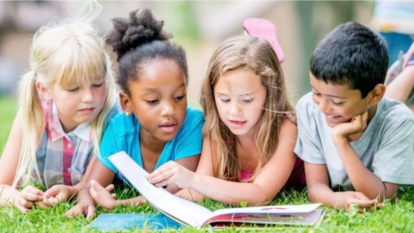 Four children of diverse ethnicities lying on grass outdoors, looking at an open book or magazine.