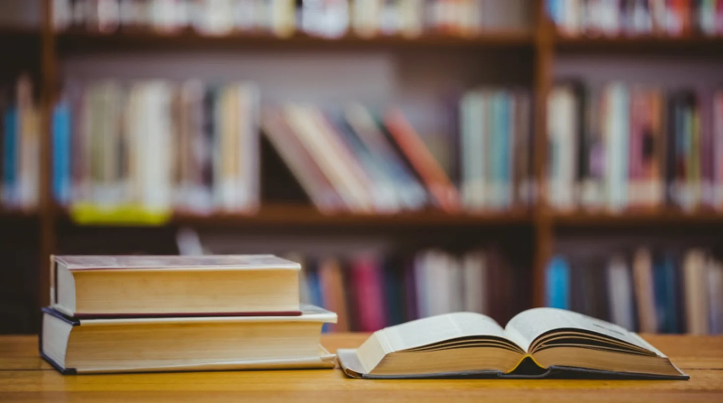 Open book and stack of three closed books on a wooden table in a library with blurred bookshelves in the background.